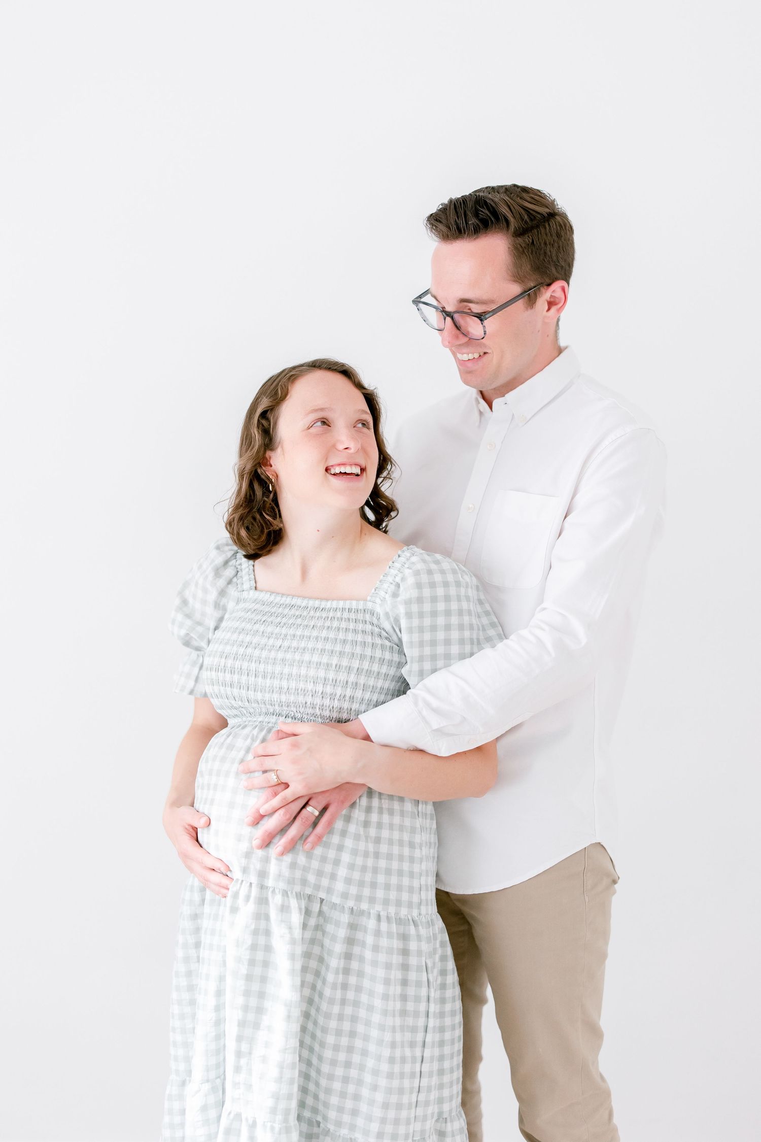 Couple in light and airy setting wearing gingham dress and white shirt embraces while laughing joyfully together.