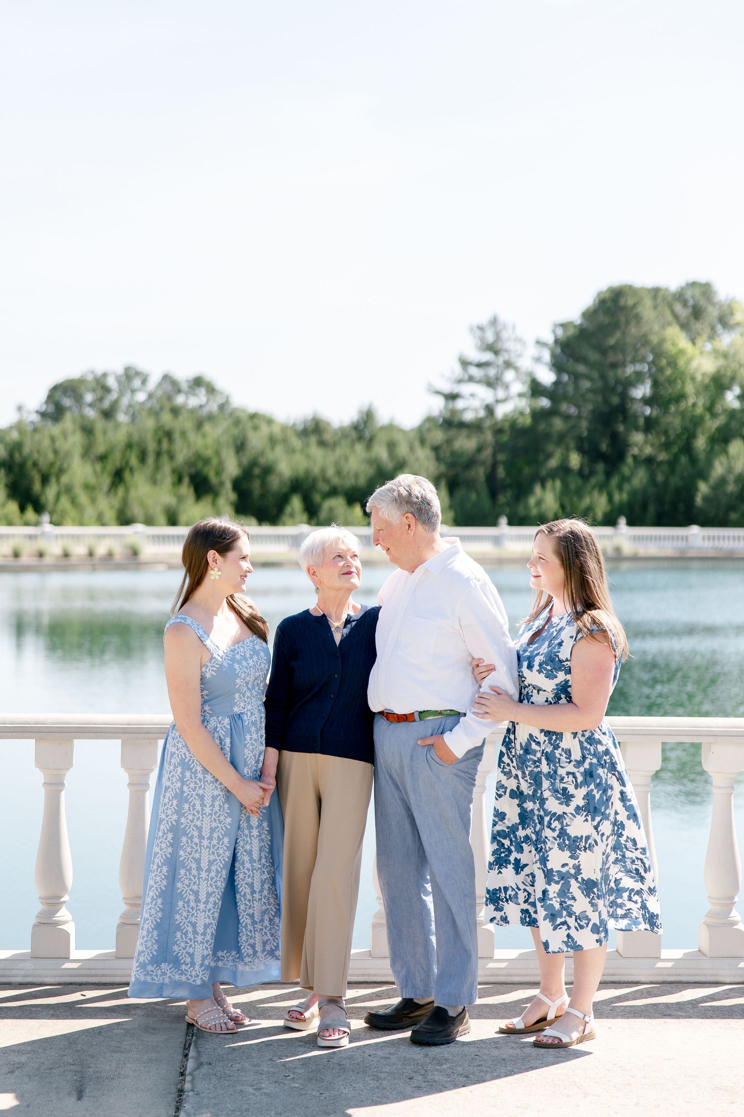 Two people in blue floral dresses and two in neutral attire share a happy moment by a scenic lake with stone balustrade.