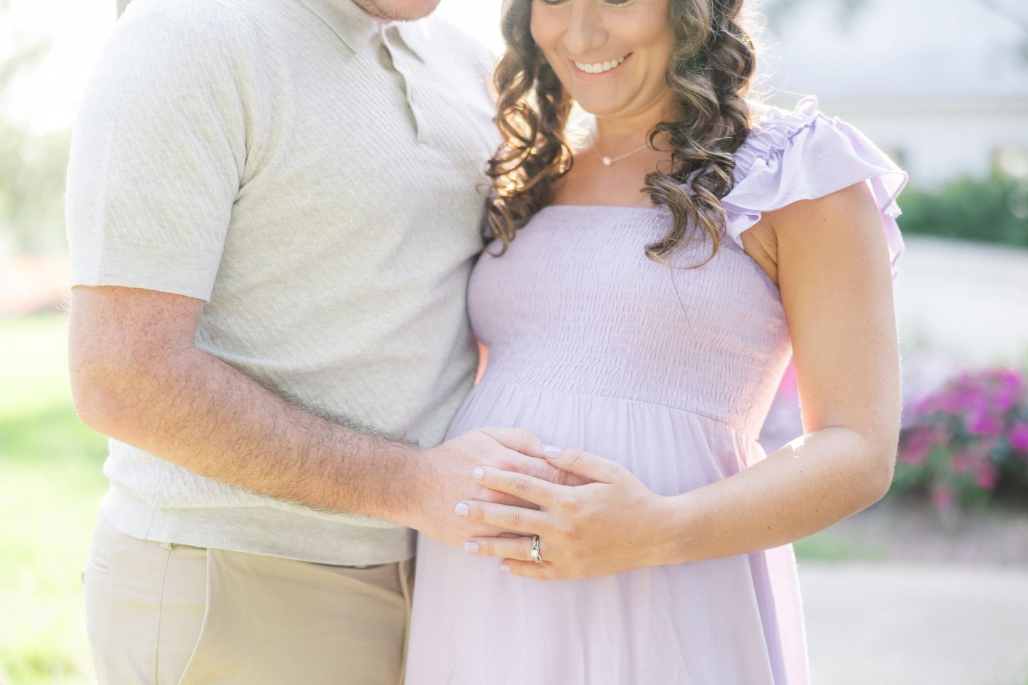 A couple wearing light-colored clothing embraces in an outdoor setting with blurred floral background.