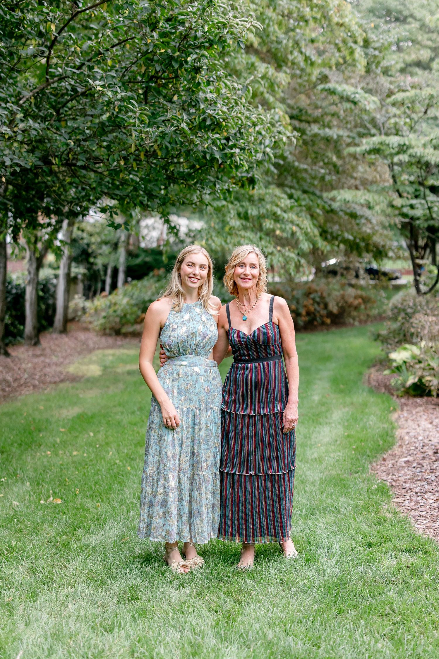 Two people wearing floral and striped dresses pose together in a lush garden setting on a grassy lawn.