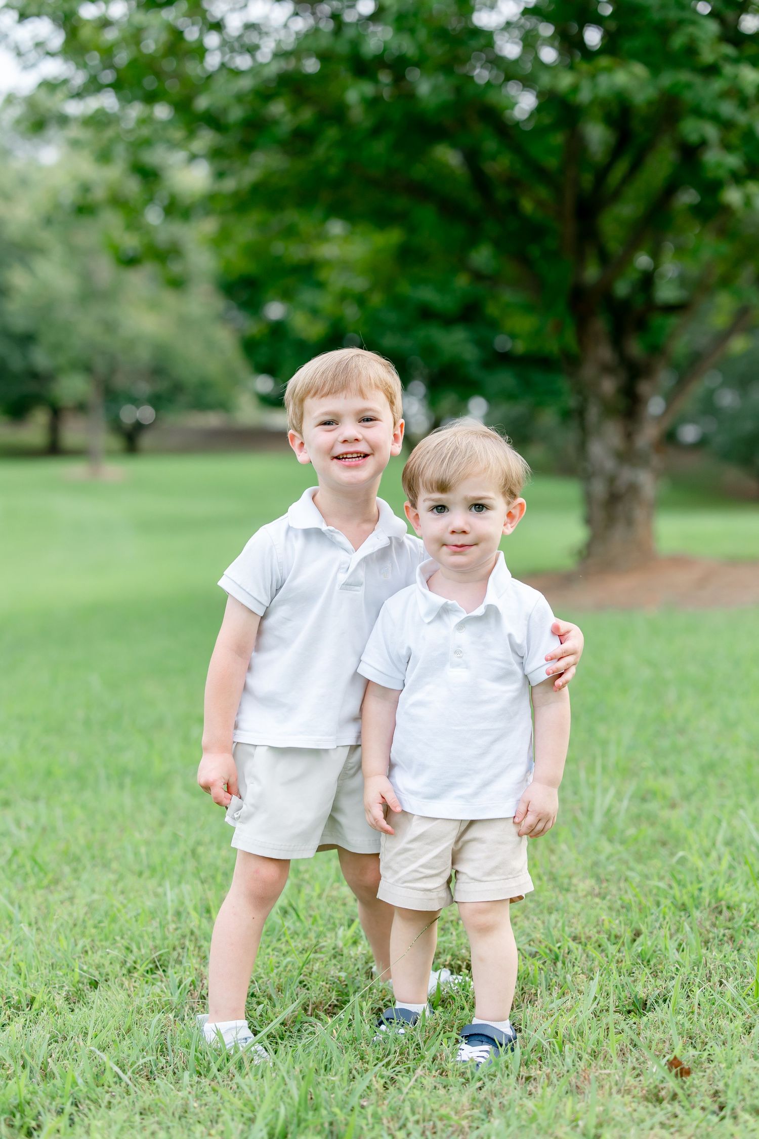 Two children in white outfits playing together on lush green grass in a park setting.