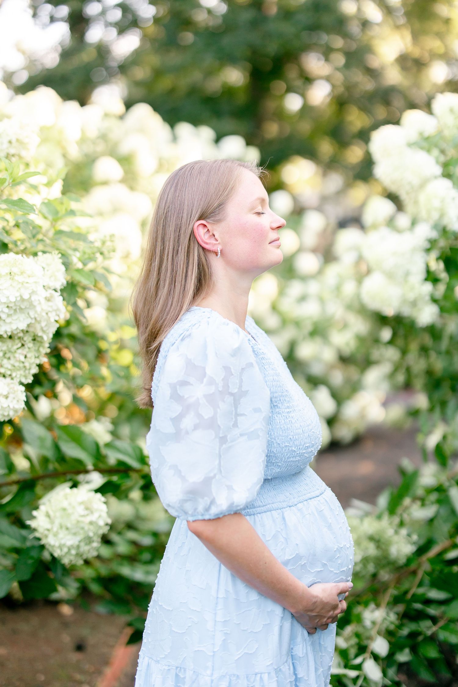 A woman in a light blue dress poses against backdrop of flowering bushes in a summer garden.