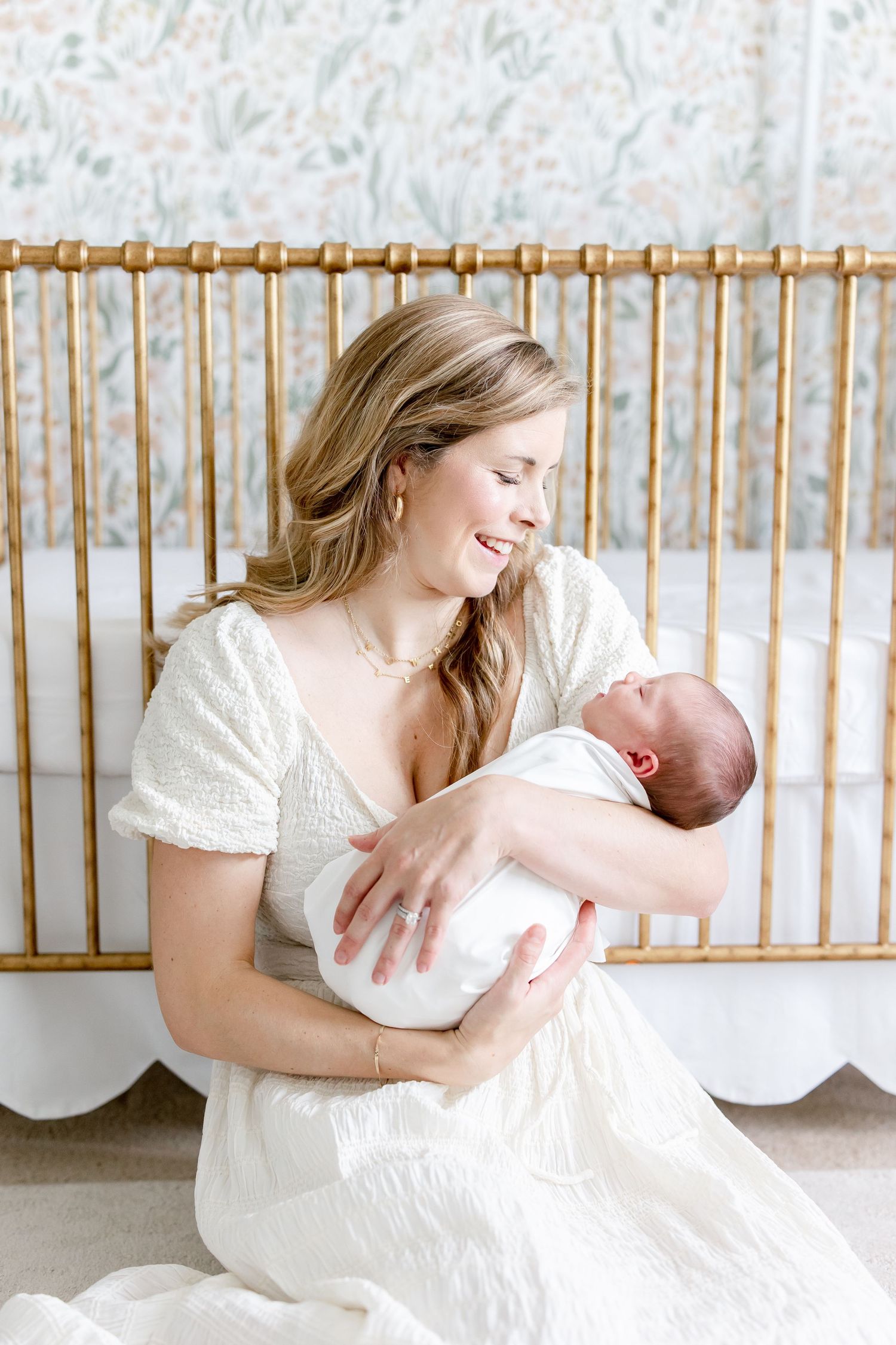 Sweet moments between mother and baby next to gold crib with floral wallpaper background.