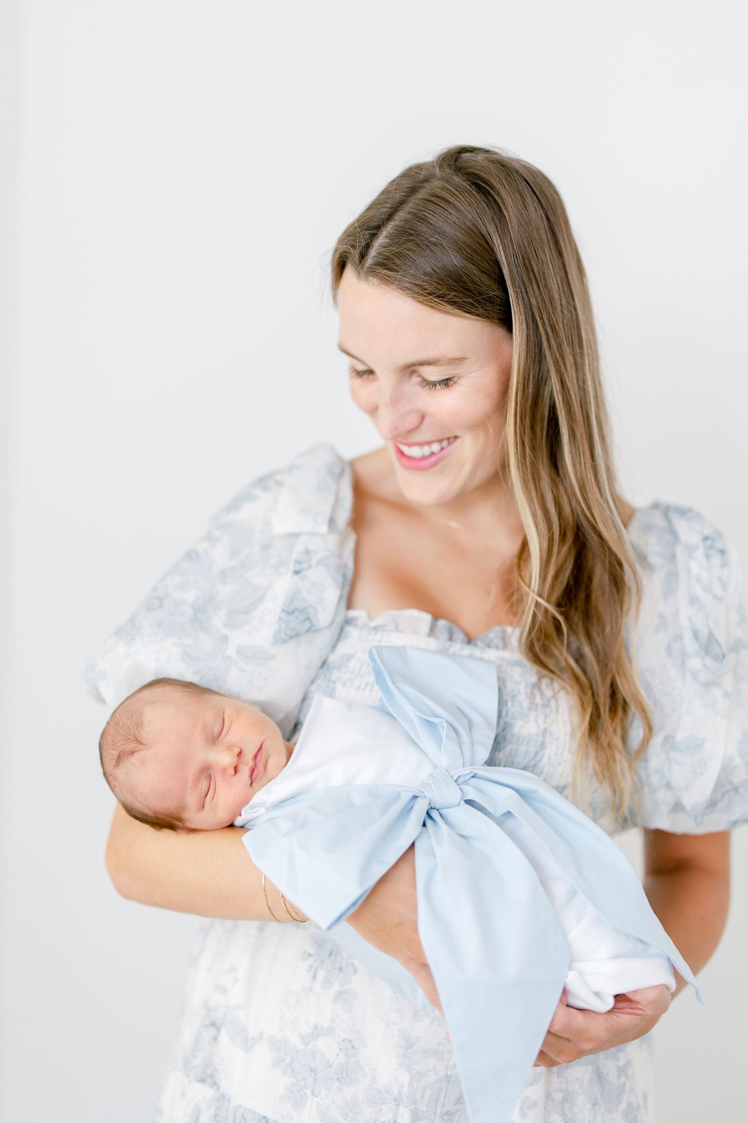 Mother in floral dress tenderly holds sleeping newborn baby wrapped in light blue swaddle blanket.