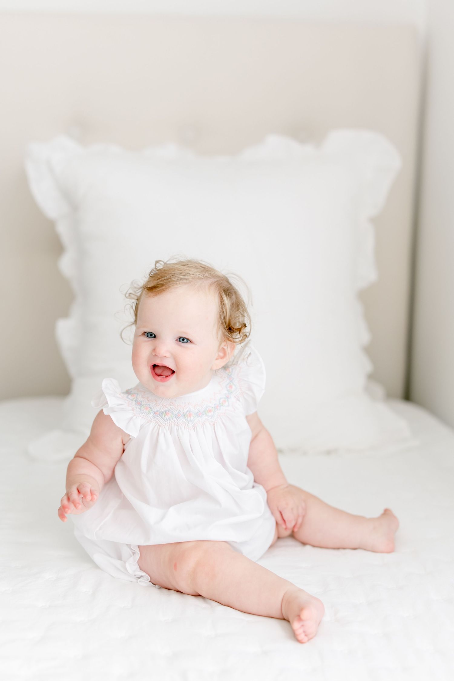 A baby in a white dress lies on a white bed in a light minimalist room against neutral walls.