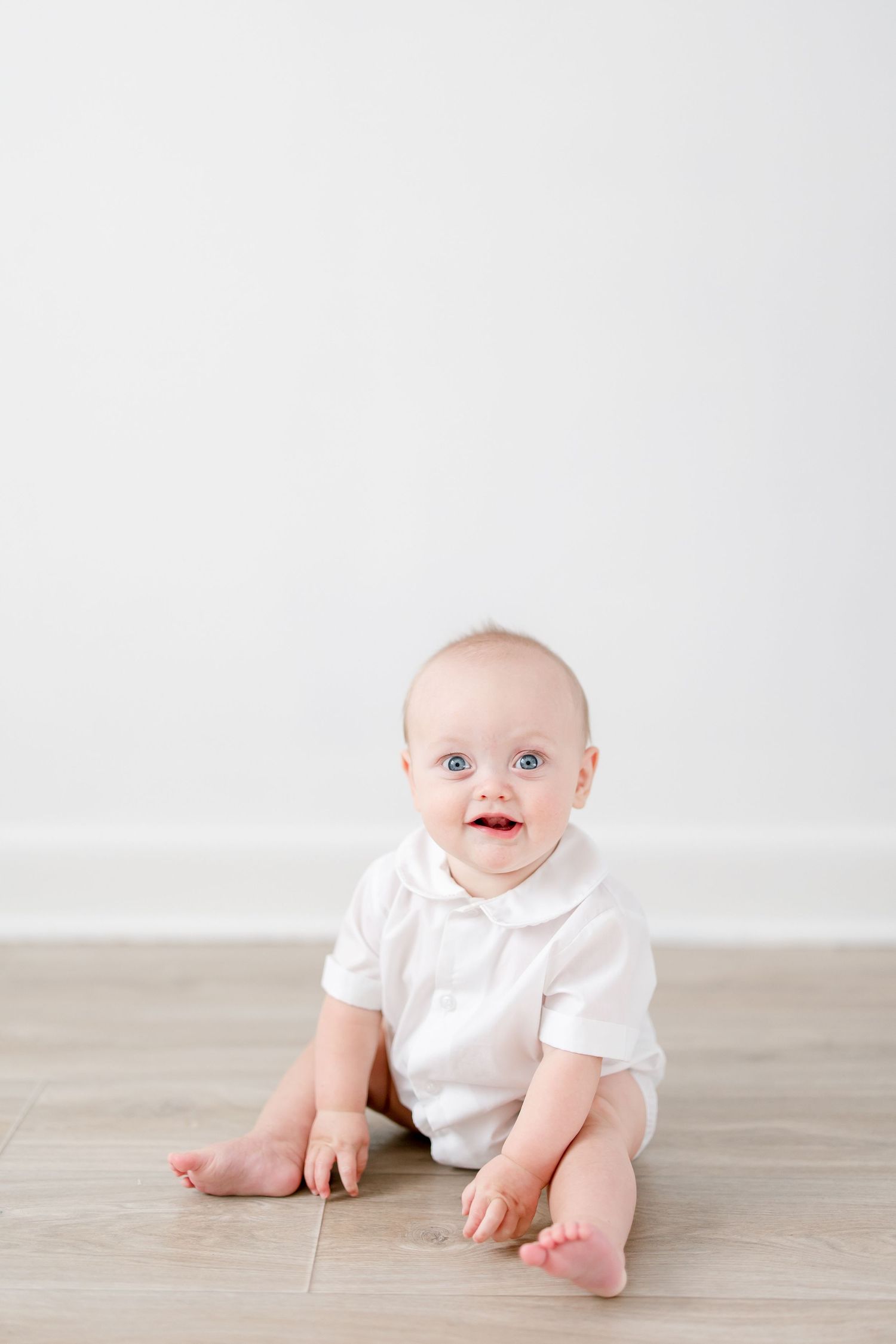 Baby in white outfit crawling on light wooden floor against white background.