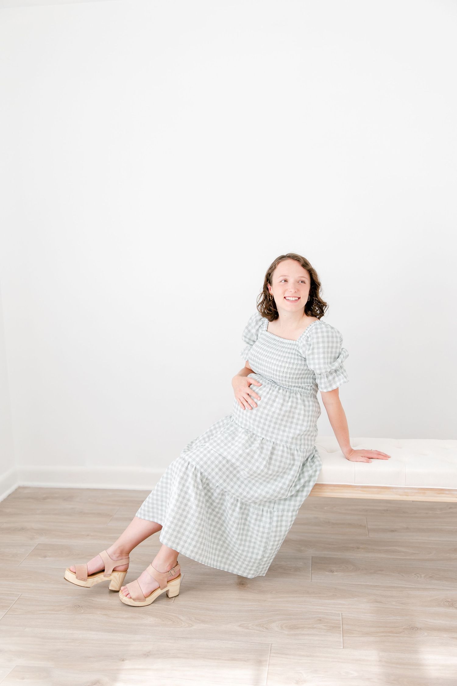 A person wearing a white and gray gingham dress poses while sitting casually on a white surface in a bright room.