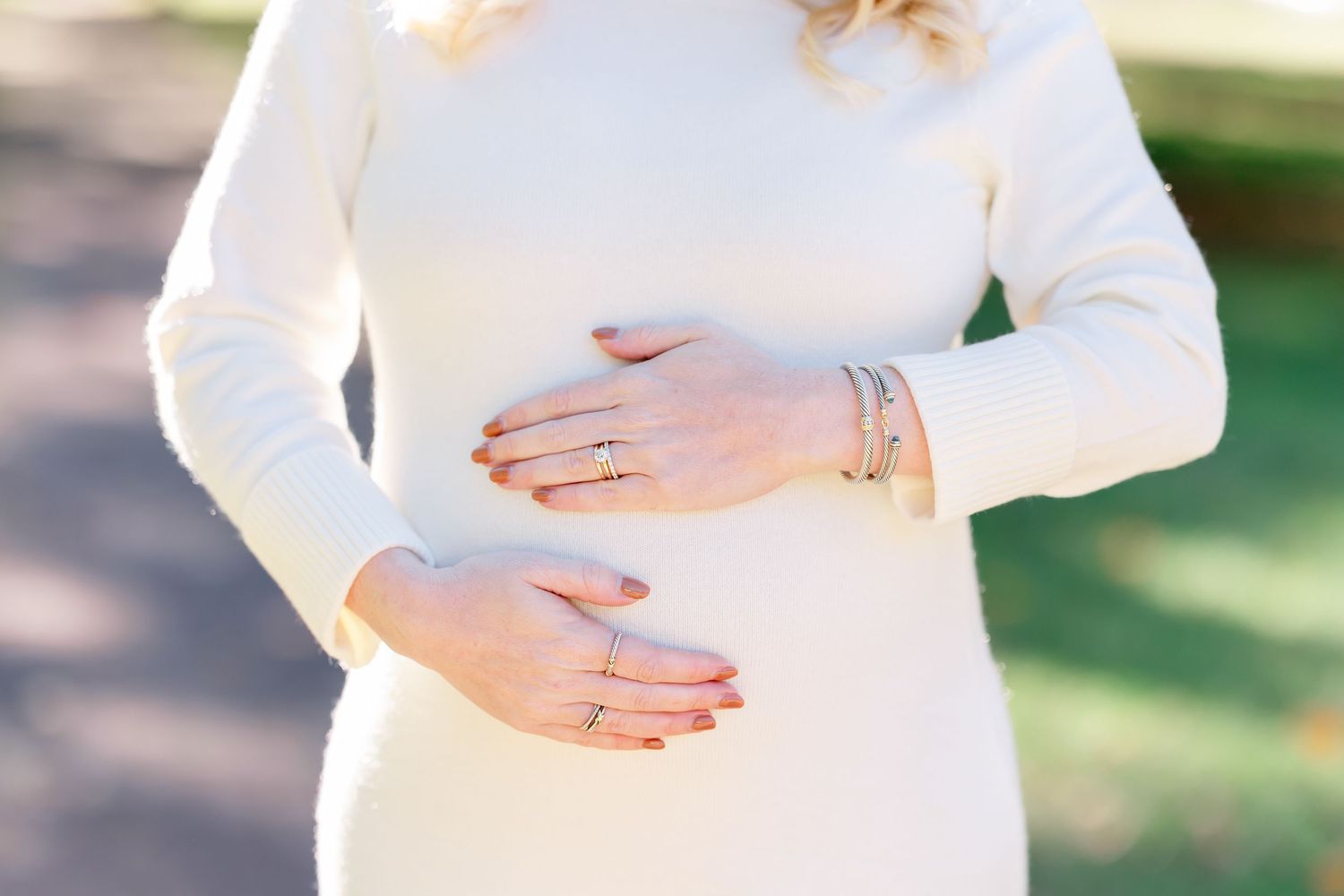 Pregnant woman in a cream knit dress gently cradling her baby bump with both hands outdoors, wearing rings and bracelets.