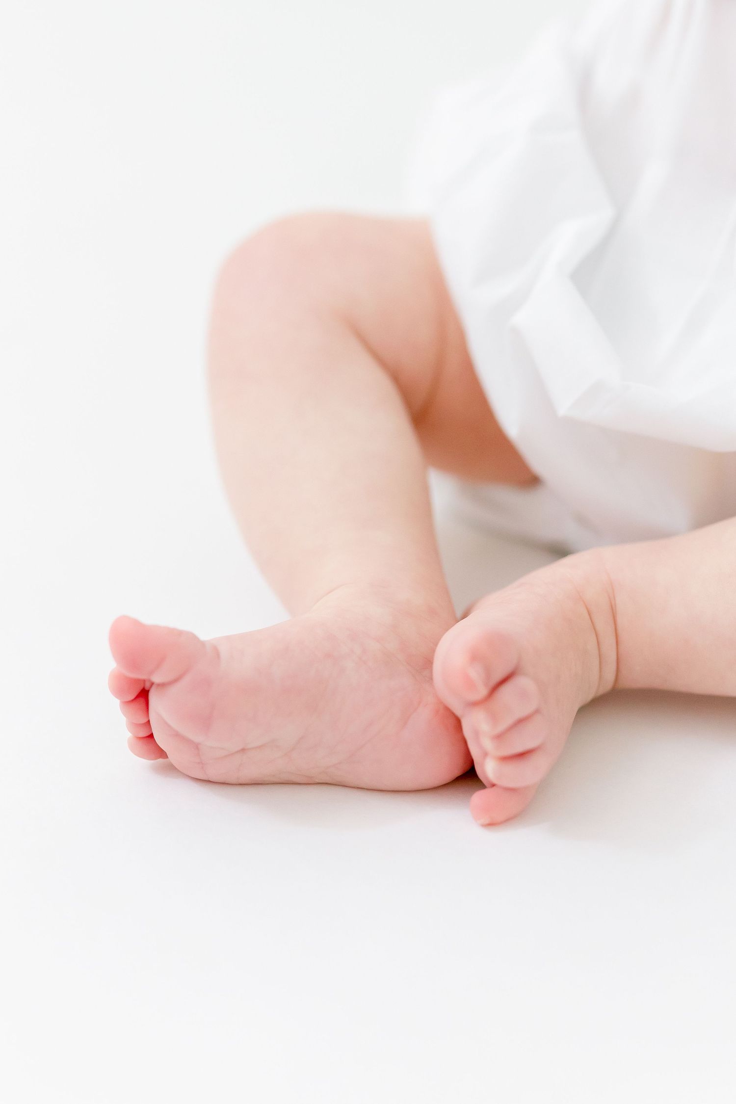 Close-up of a newborn baby's tiny bare feet and chubby legs against a soft white background, wearing a white outfit.