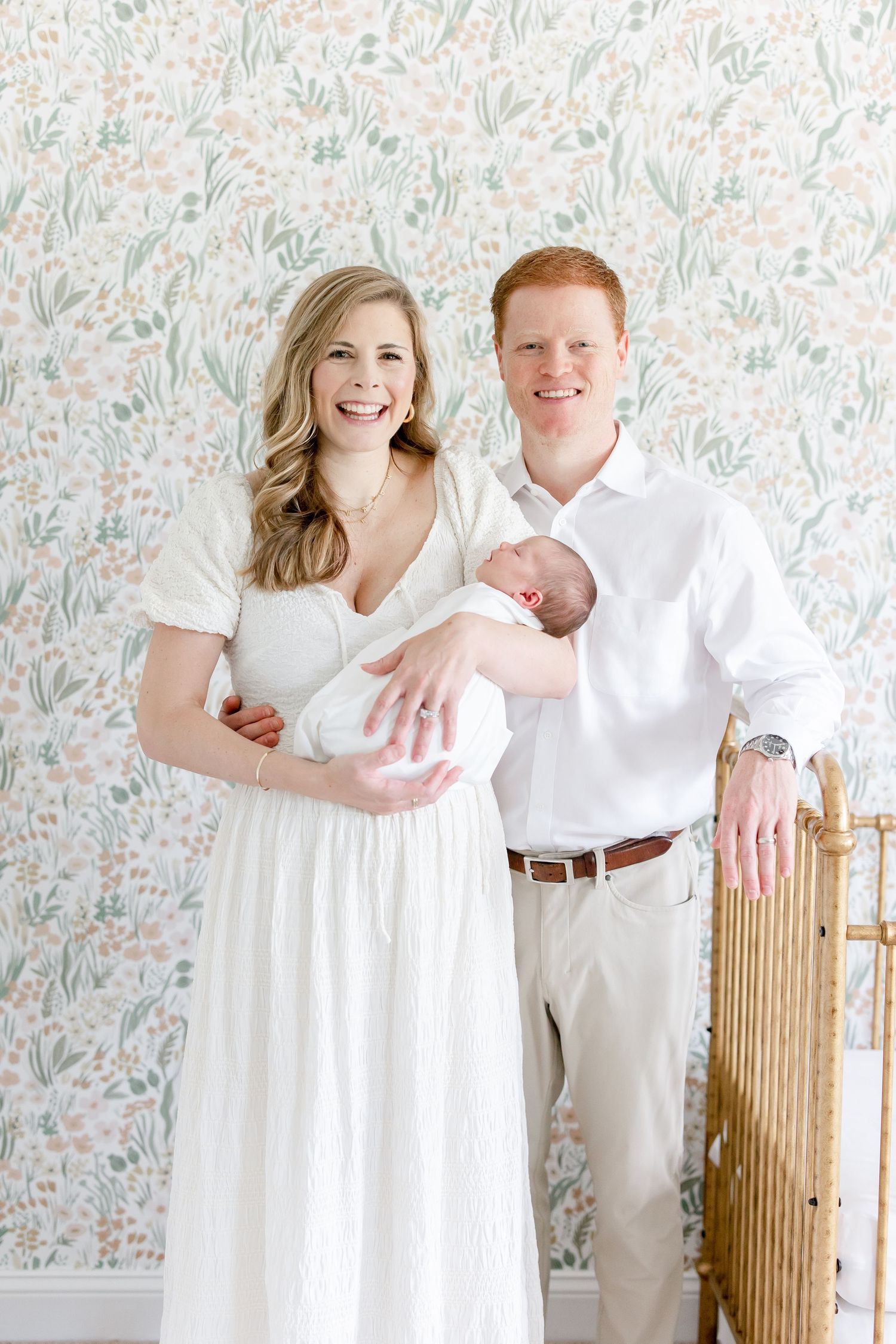 A couple in white clothing poses with their newborn against floral wallpaper.