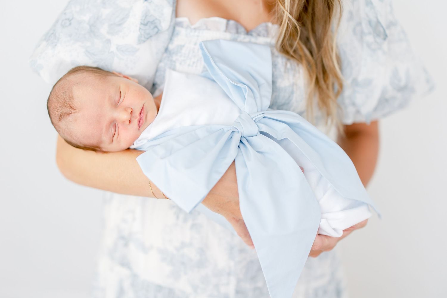 A sleeping newborn baby wrapped in a light blue bow is held gently against someone wearing a white lace dress.