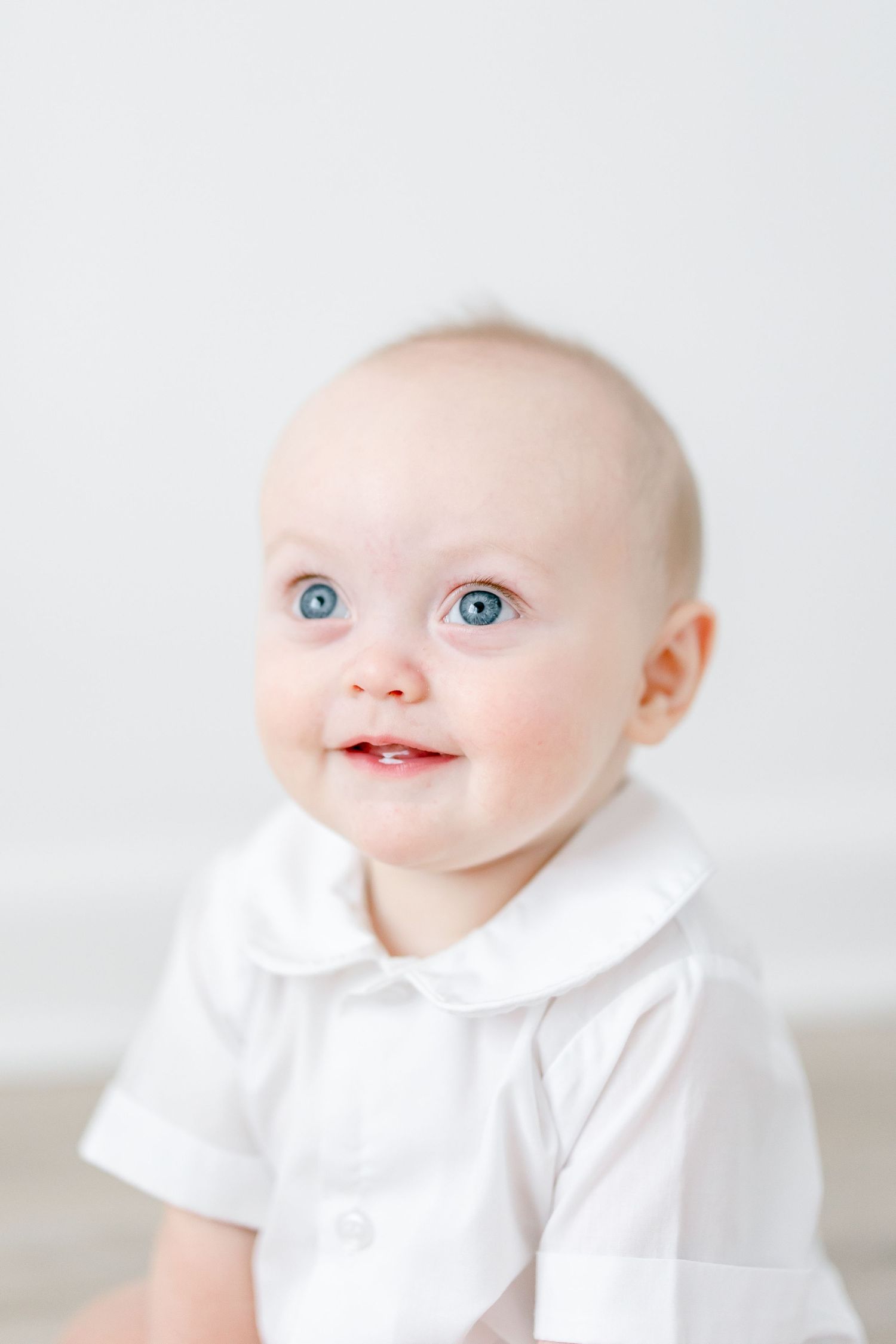 Baby in white collared outfit smiling brightly against minimal white background.