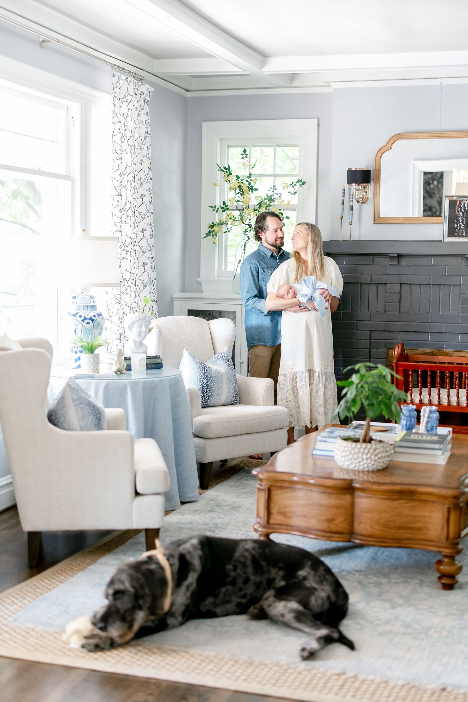 Couple holding newborn in bright living room with sleeping dog on rug and coastal decor.