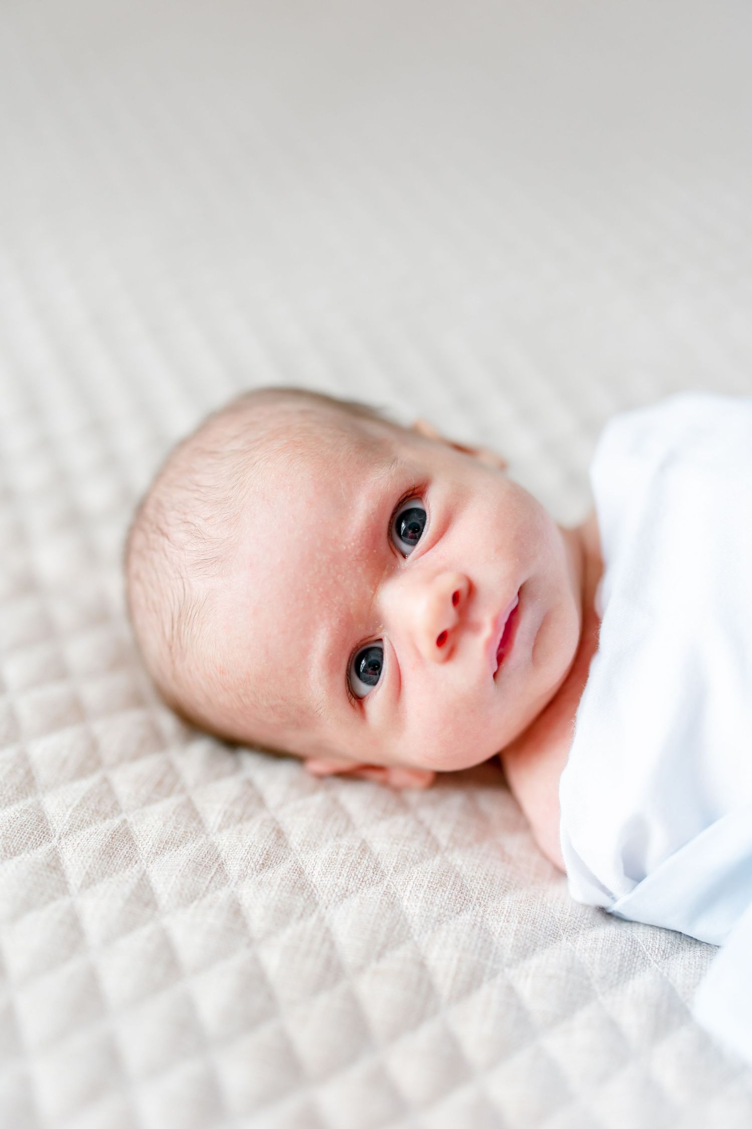 Sweet baby with bright eyes lies peacefully on white textured bedding while looking at the camera.