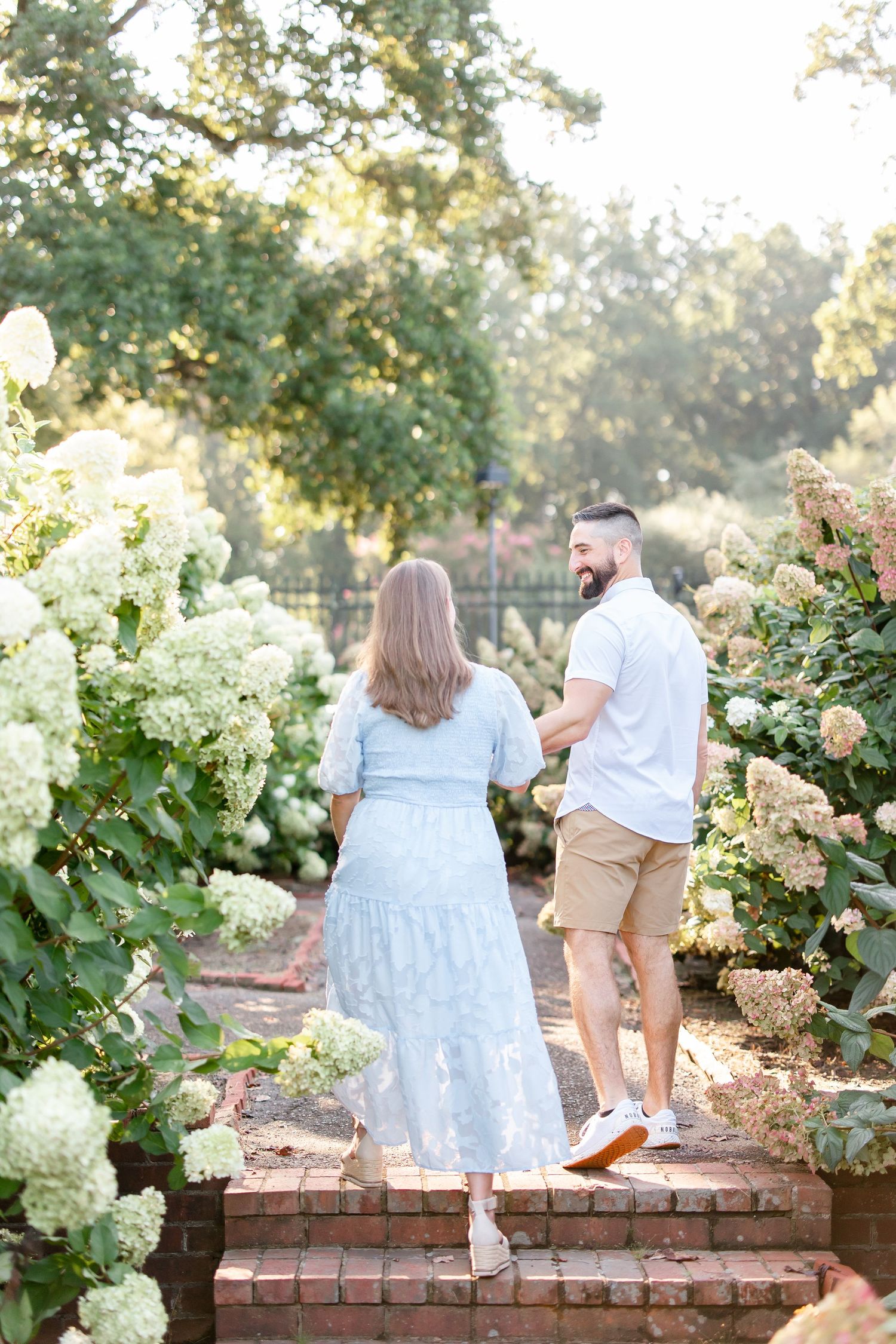 Couple sits together on brick garden wall surrounded by blooming white hydrangeas.