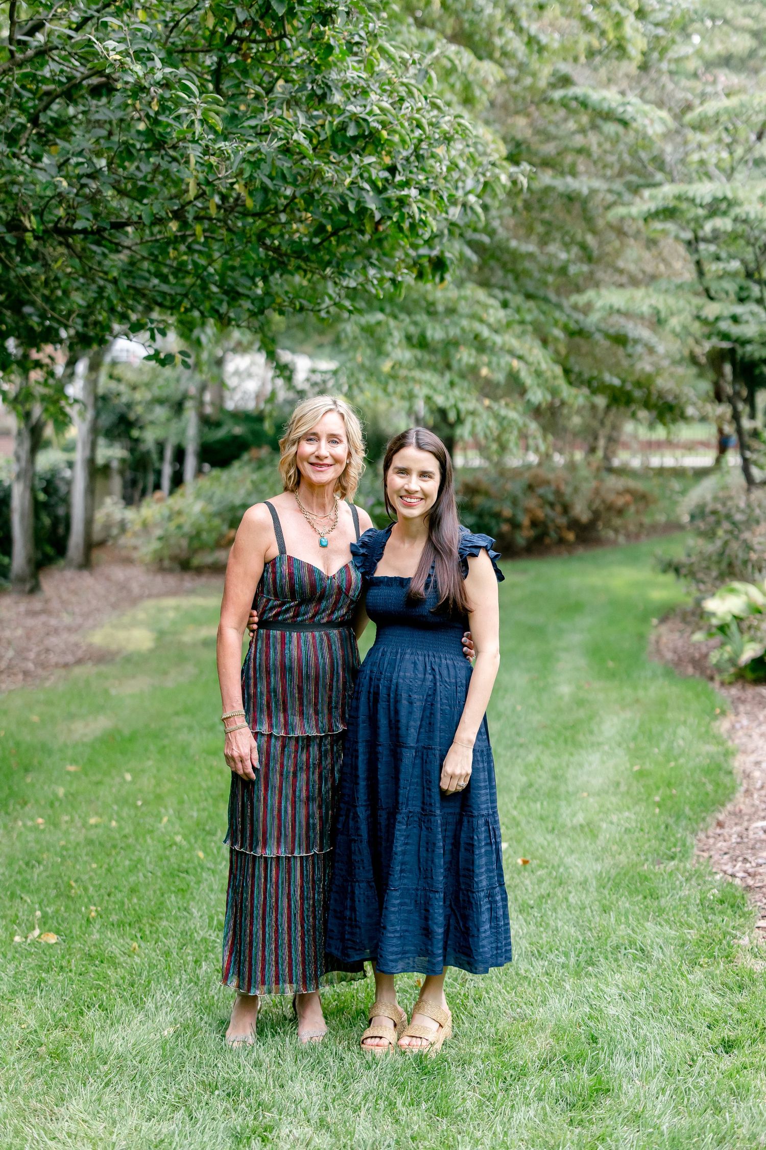 Two people in summer dresses stand together on a grassy lawn surrounded by green trees.