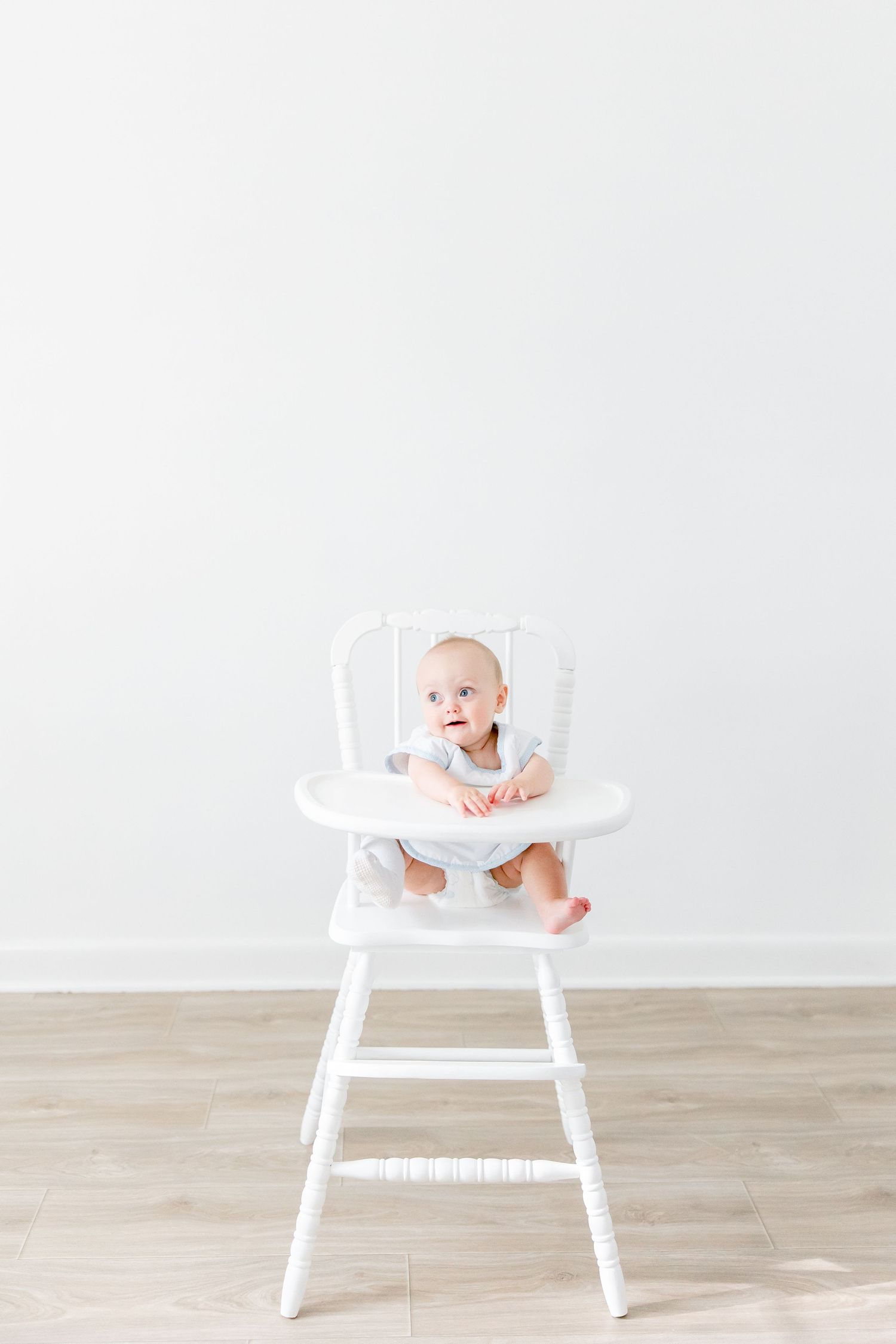 Baby sitting in white wooden high chair against minimalist white wall and wood floor.