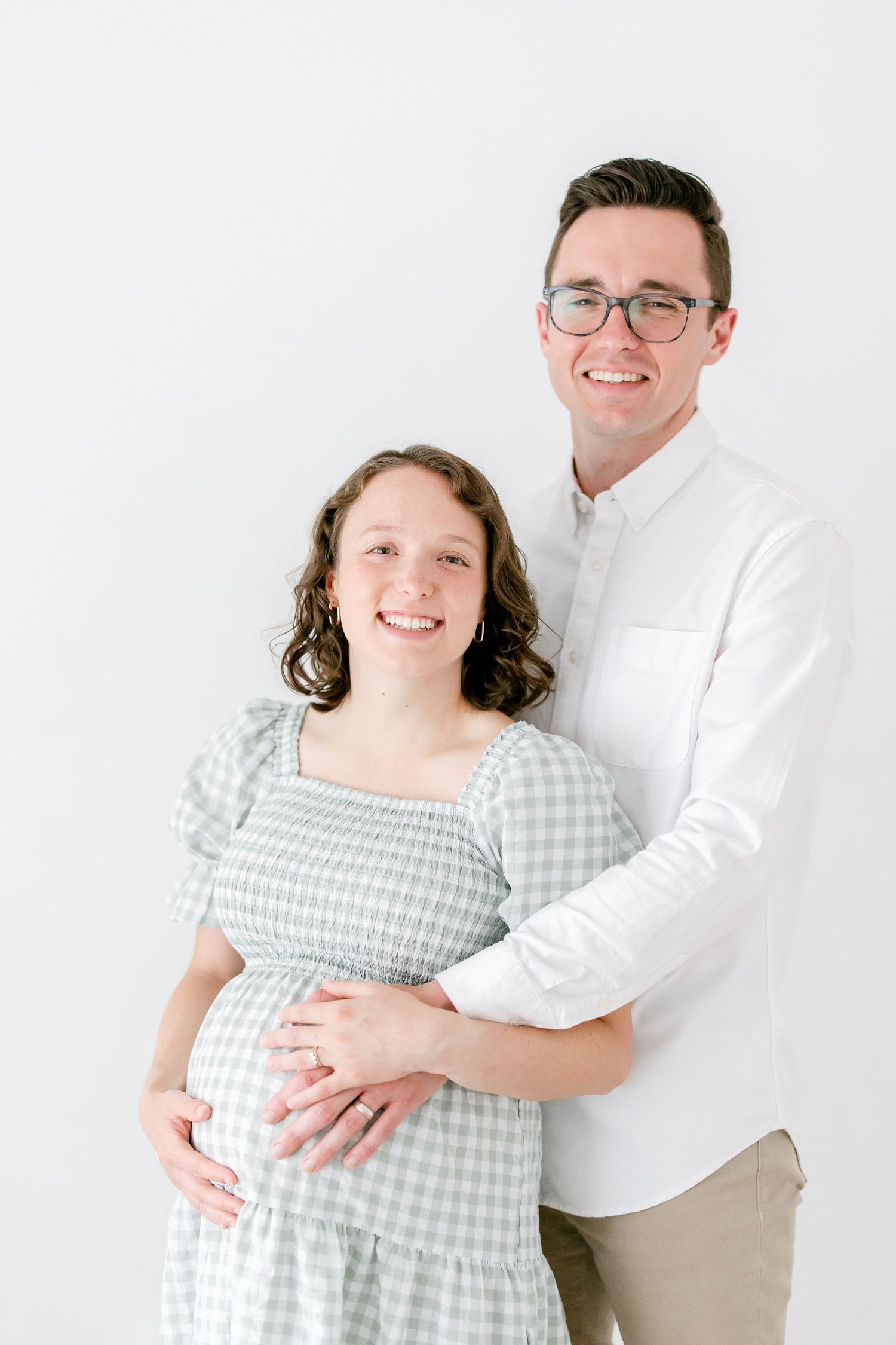 A couple poses together against a white backdrop, with the woman wearing a gray gingham dress and the man in a white shirt.