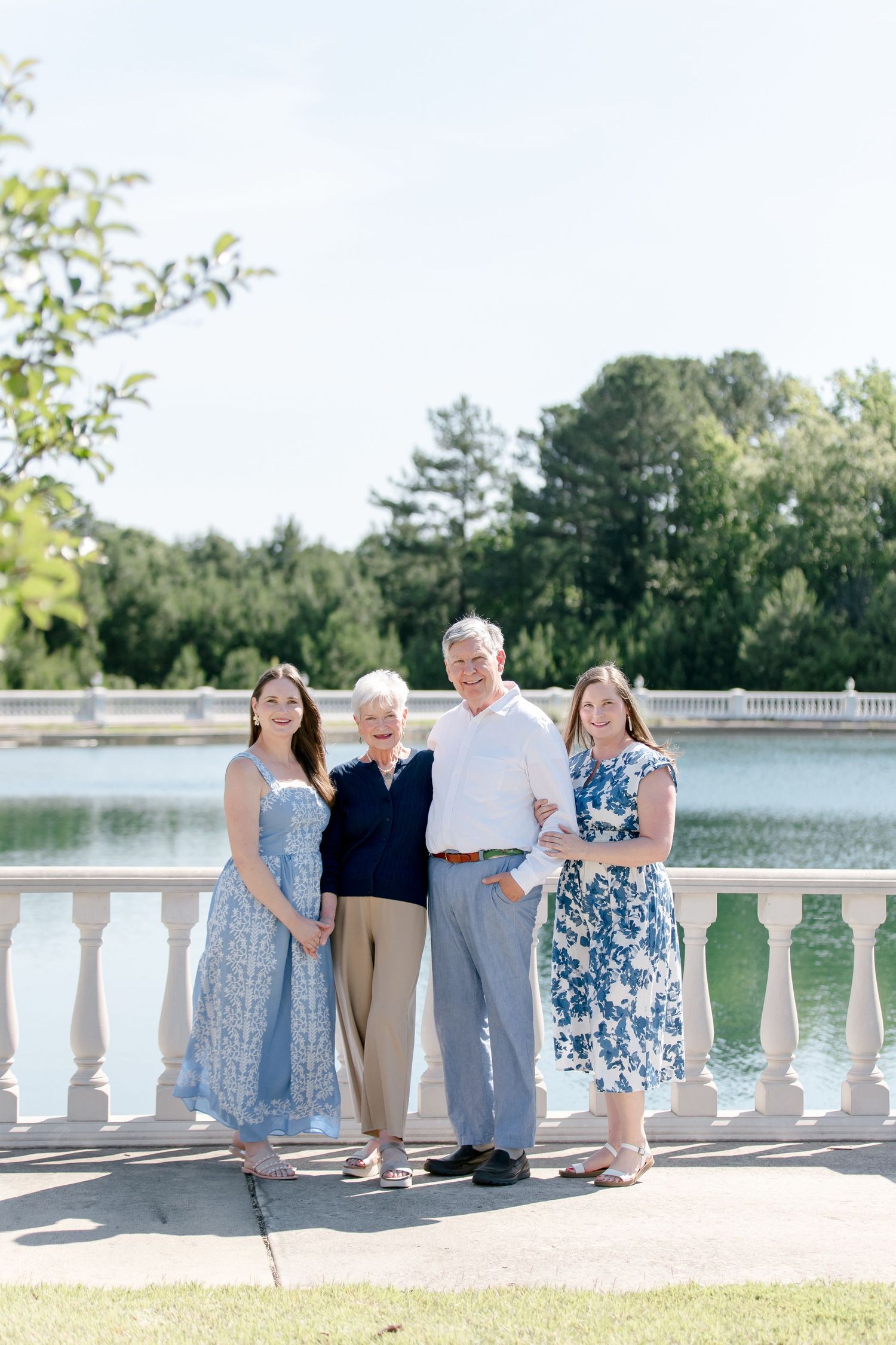 A group poses together on a white stone balcony overlooking a serene lake surrounded by trees on a sunny day.