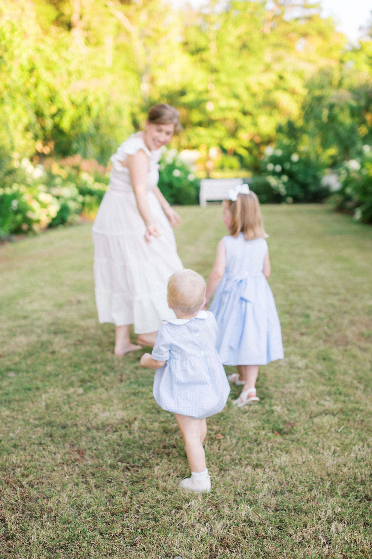 Three figures in light dresses walking together through a sunlit garden.