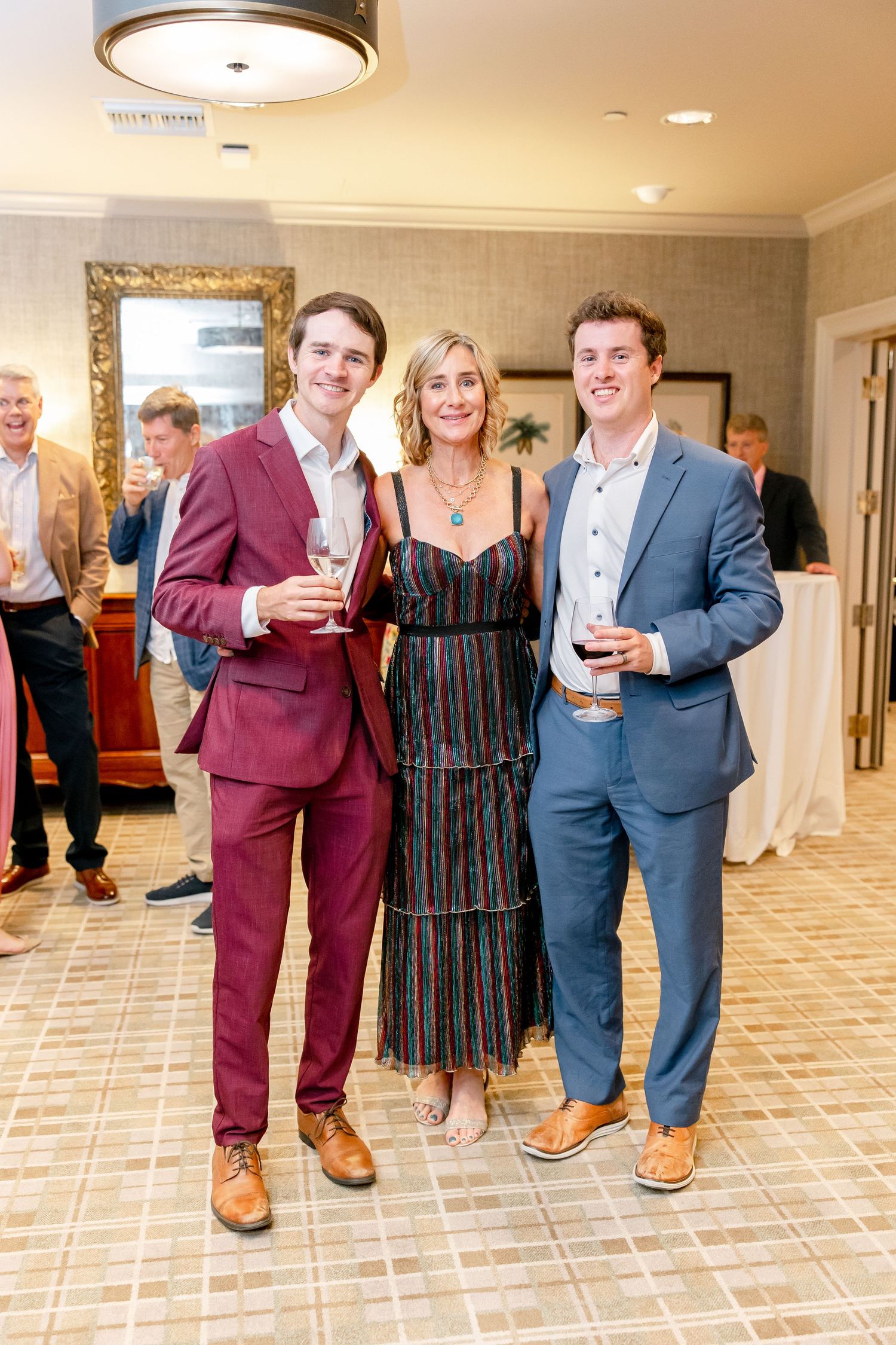 Three people in formal attire pose together at an elegant event with a decorative gold-framed mirror in background.