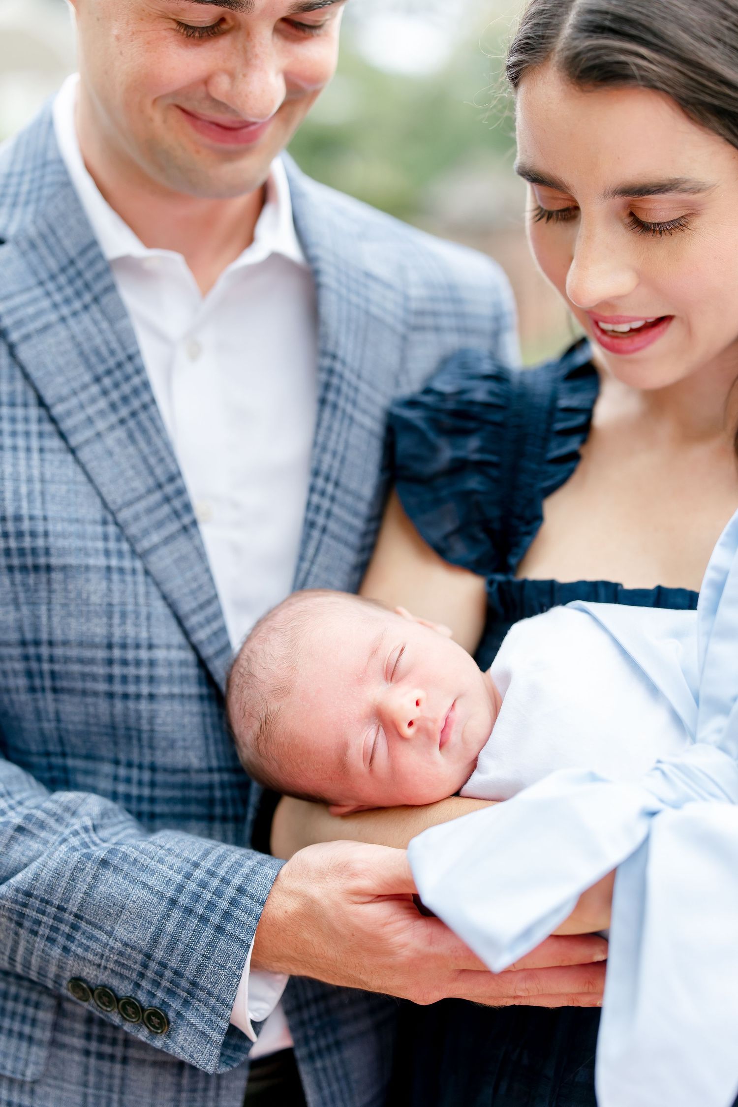 Couple in outdoors setting holding and admiring newborn baby wrapped in white.