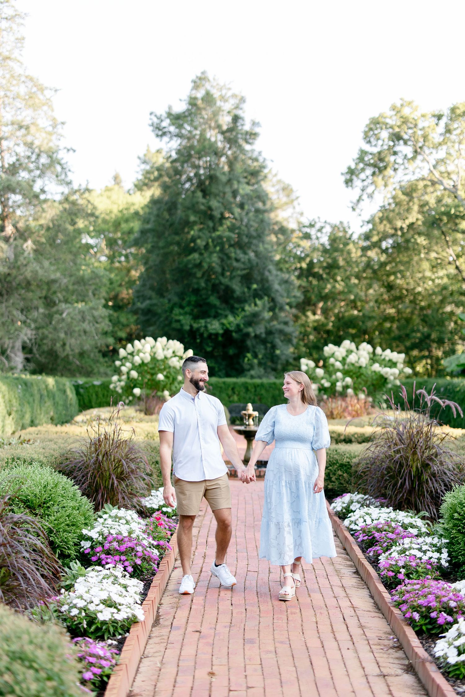 A couple strolls along a brick pathway surrounded by blooming pink flowers and lush garden landscaping in an outdoor setting.