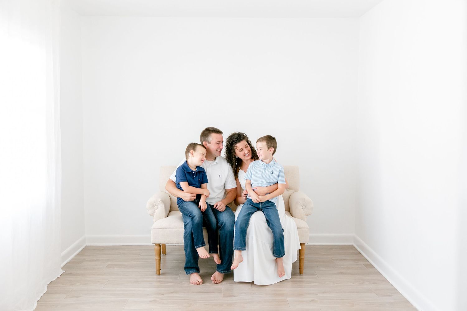Family with three children dressed in blue and white sits on cream couch in white room during photo session.