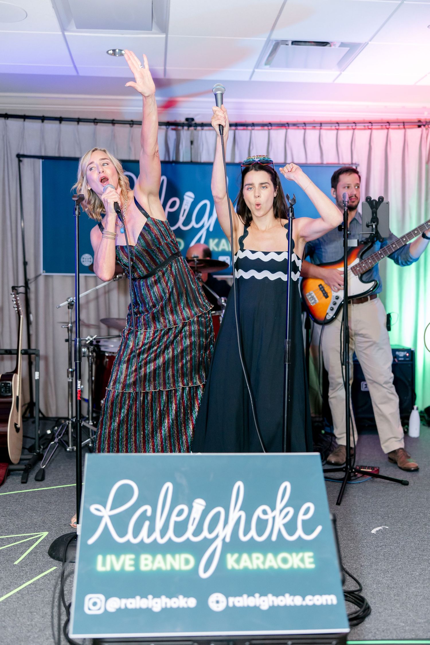 Two singers performing energetically on stage with a live band at a Raleighoke karaoke event with colored lighting.