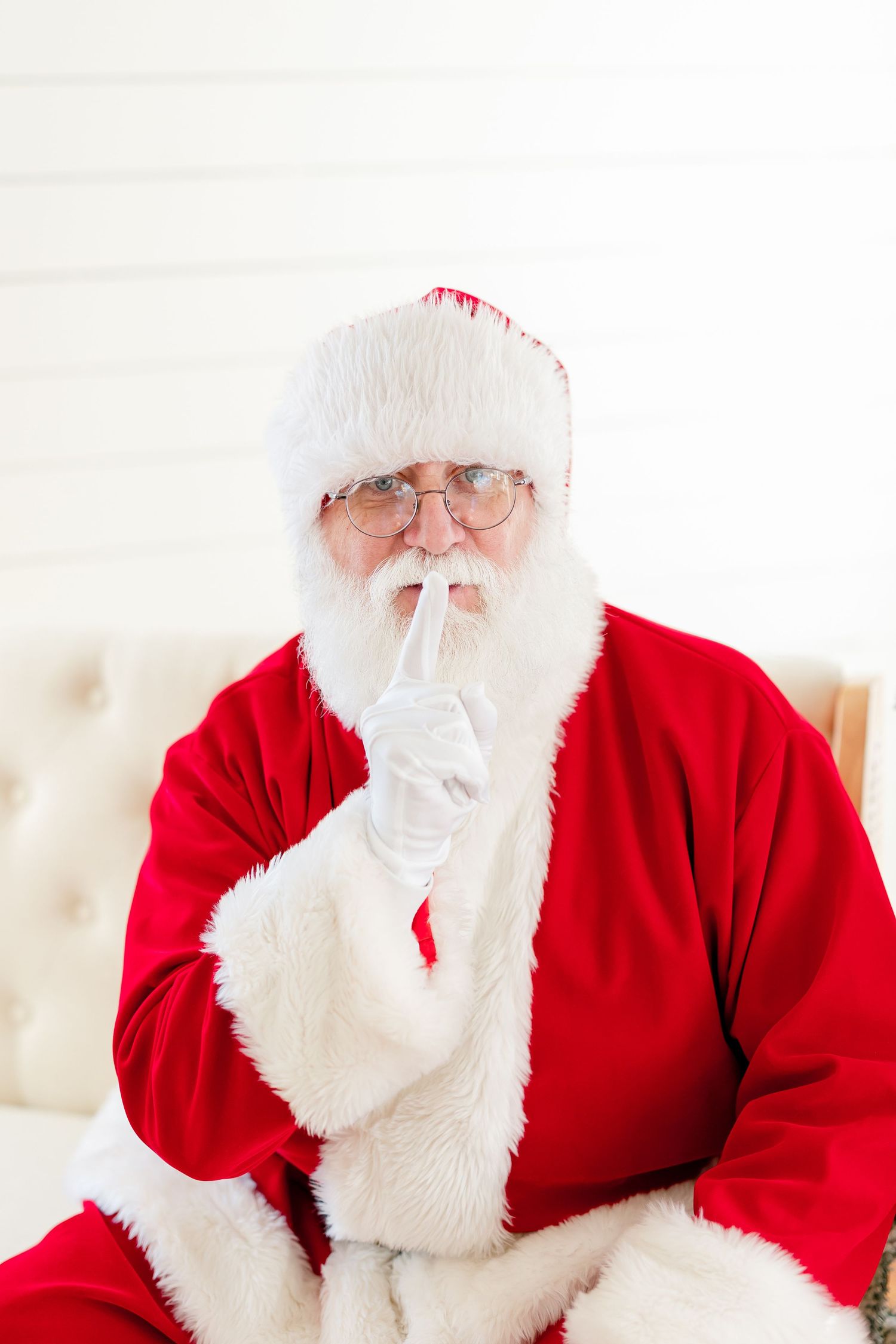 Santa Claus in red suit making a shushing gesture with finger to lips, wearing glasses and white gloves.