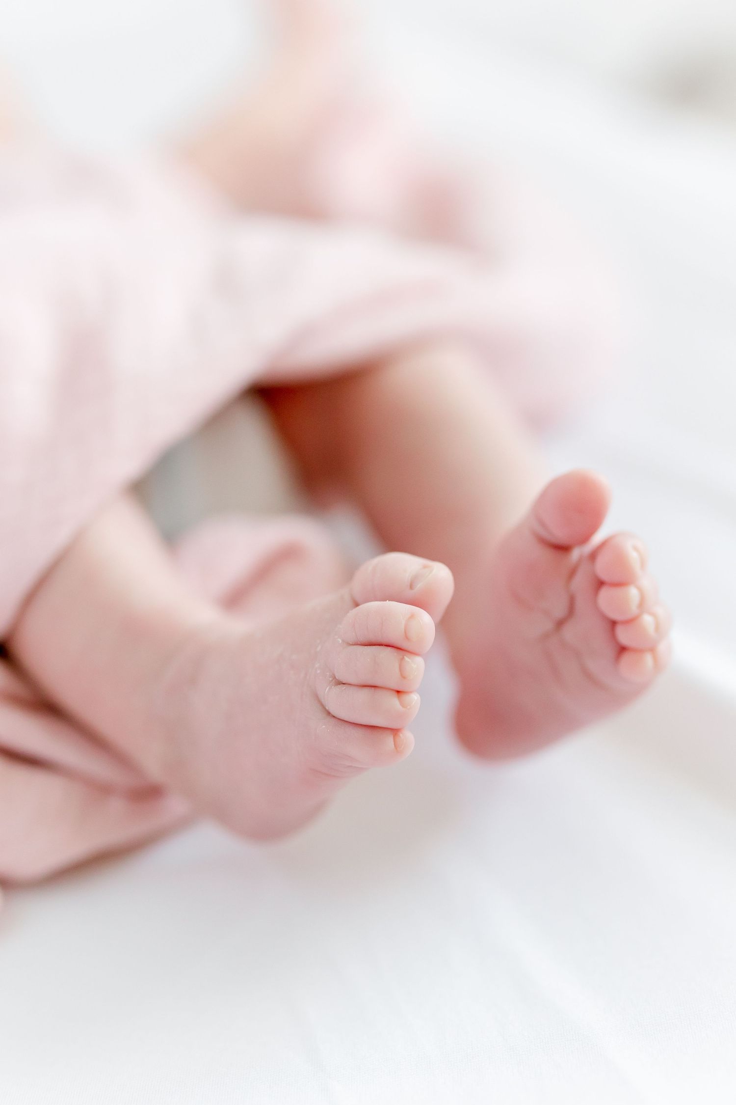 Close-up photo of tiny newborn baby feet in soft pink tones showing delicate toes and skin texture against blurred background.