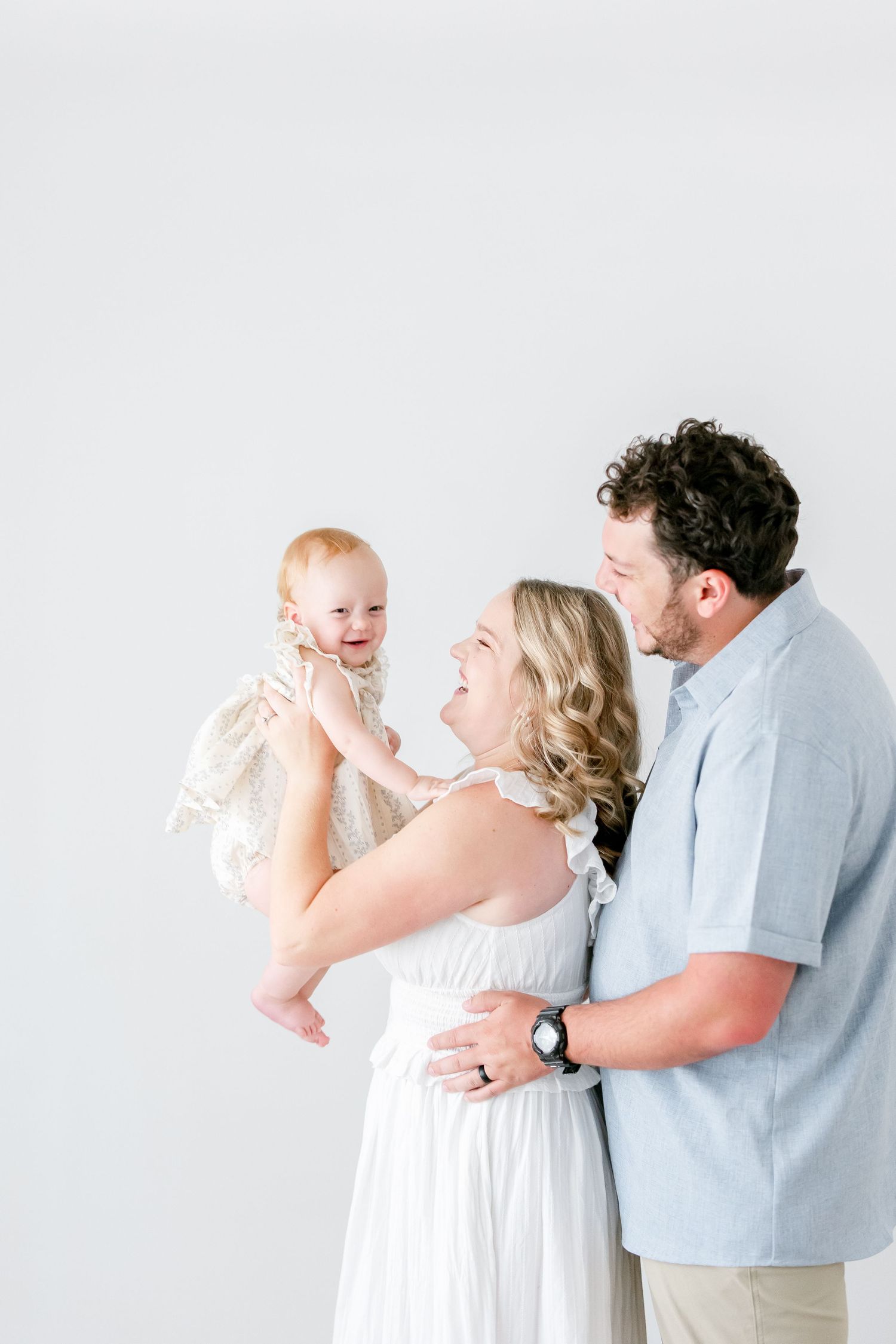 Family poses together against a white background wearing light colored clothing and sharing joyful expressions.