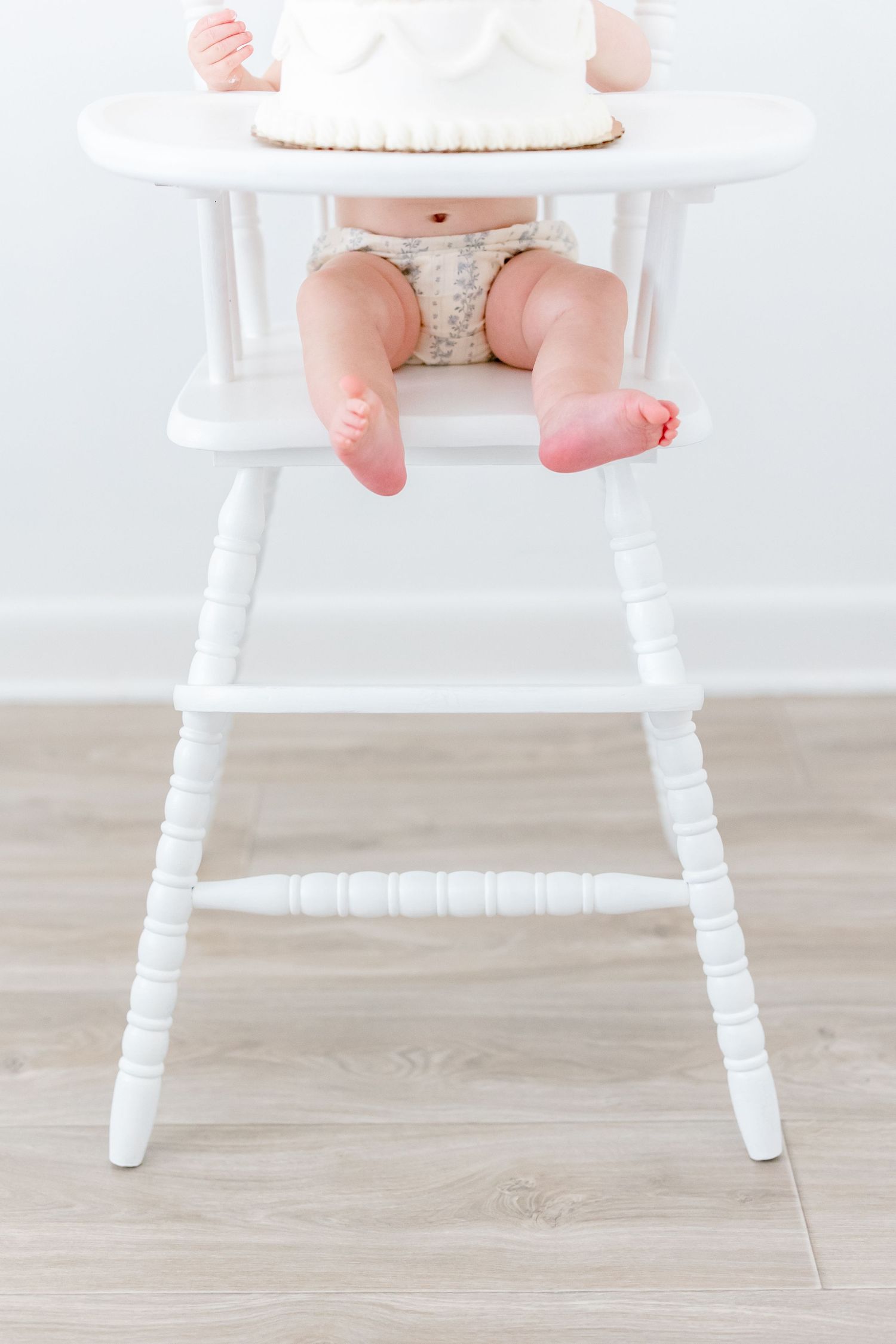 Tiny feet and legs in a diaper sitting in a white wooden high chair on light wood flooring.