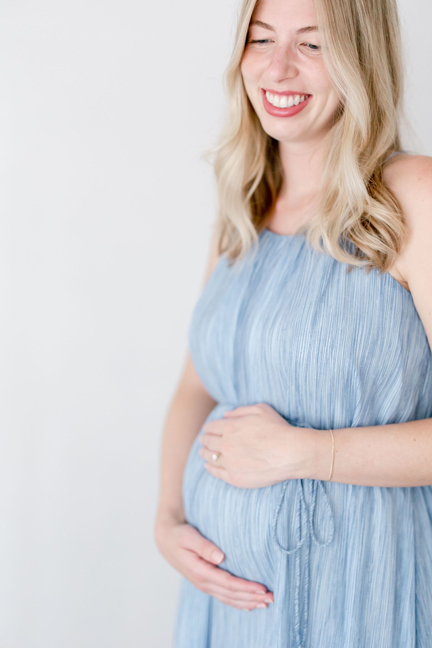Smiling woman in light blue pleated dress poses against white backdrop during pregnancy session.