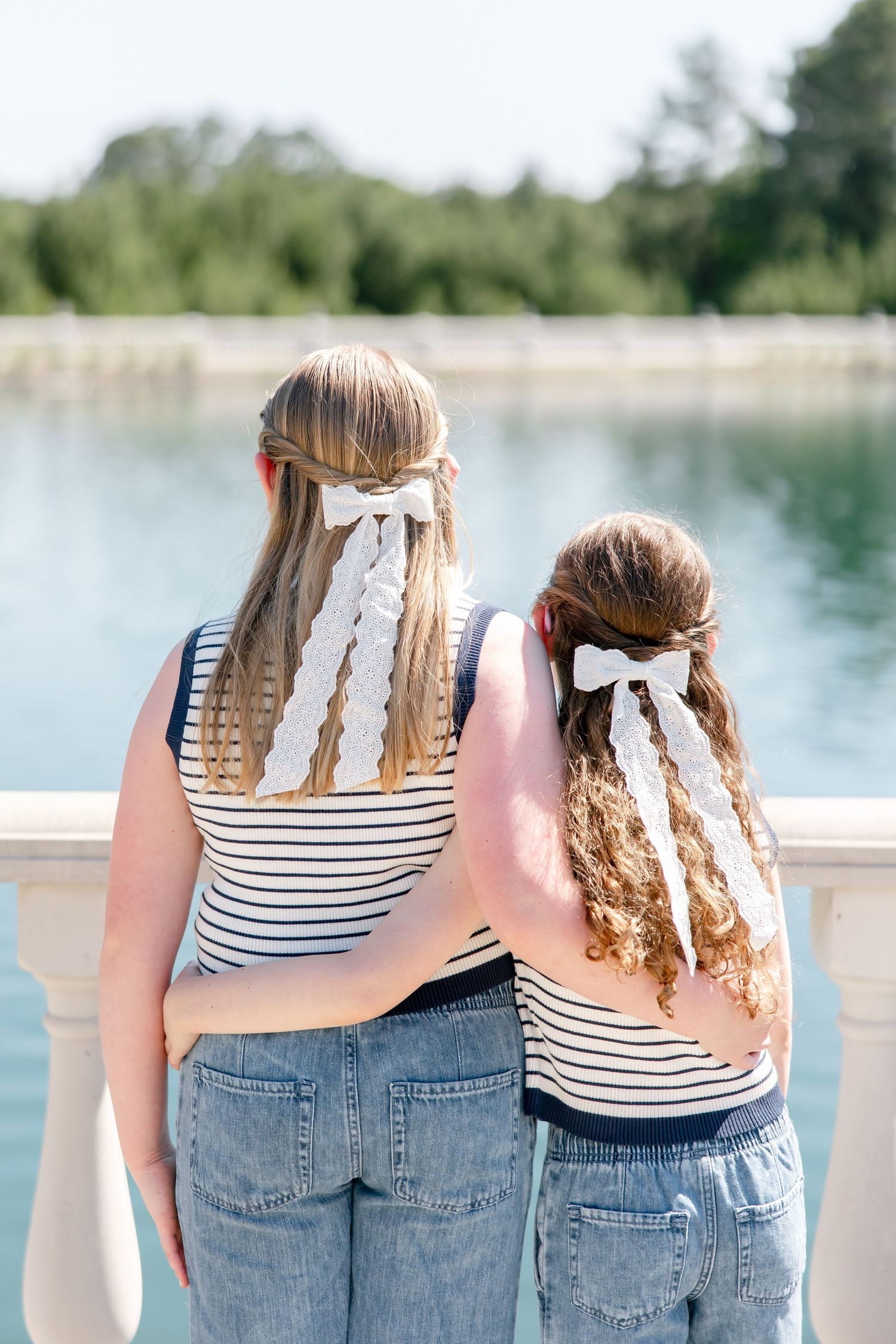 Two people in matching striped tops and jeans looking out at a lake with white ribbon hair bows.