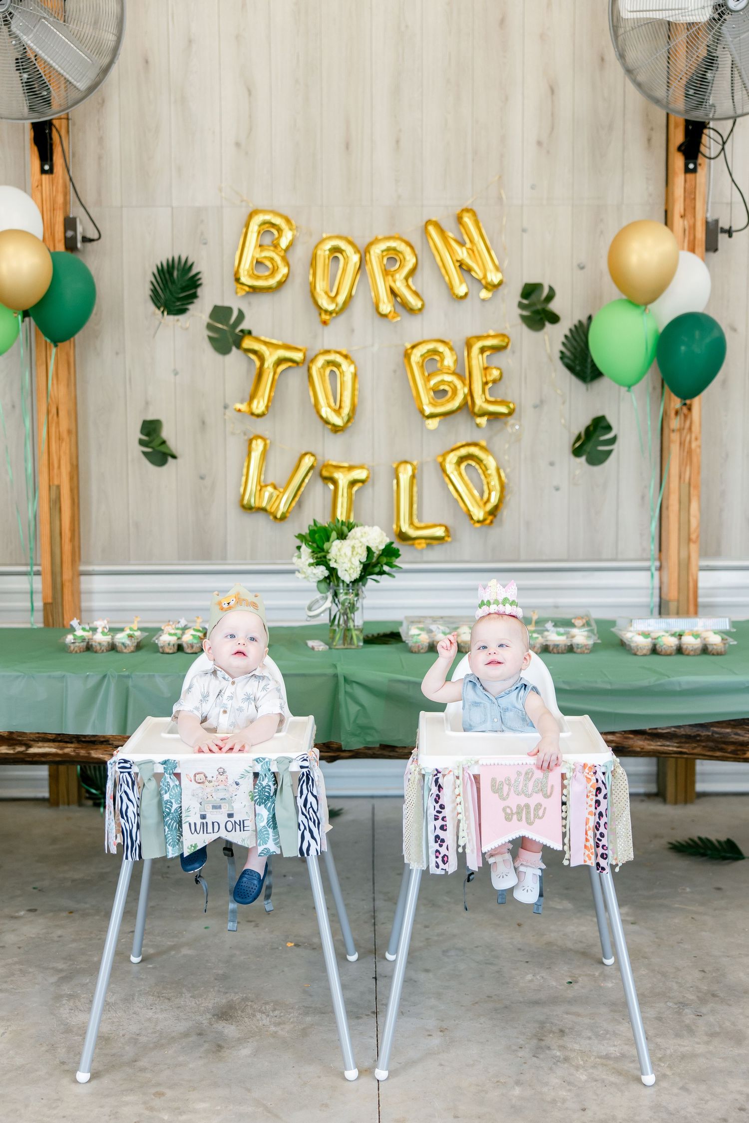 Twin babies in highchairs under 'Born to be Wild' balloon banner with party decorations.