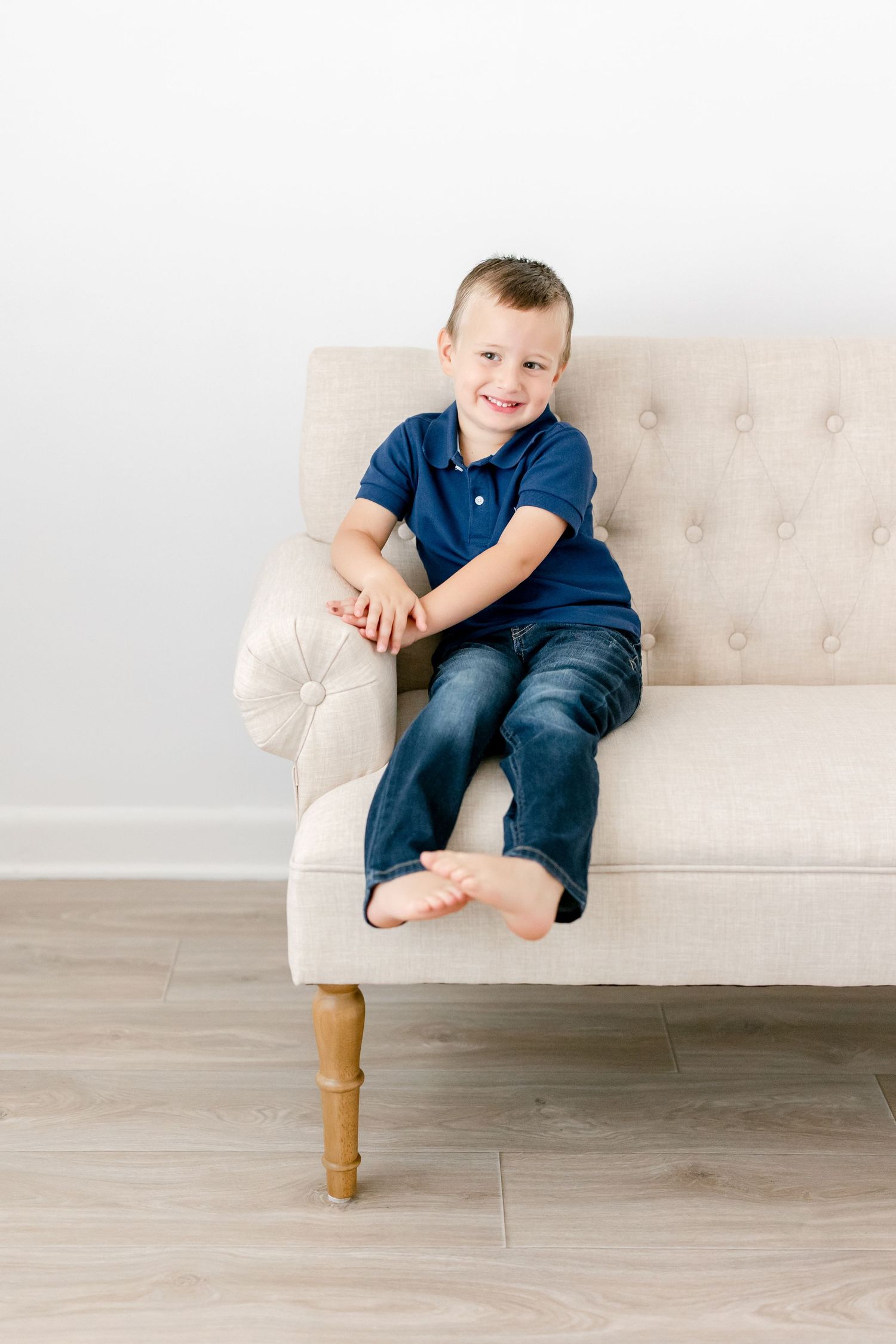 Child in navy blue shirt and jeans sits casually on beige tufted sofa against white background.