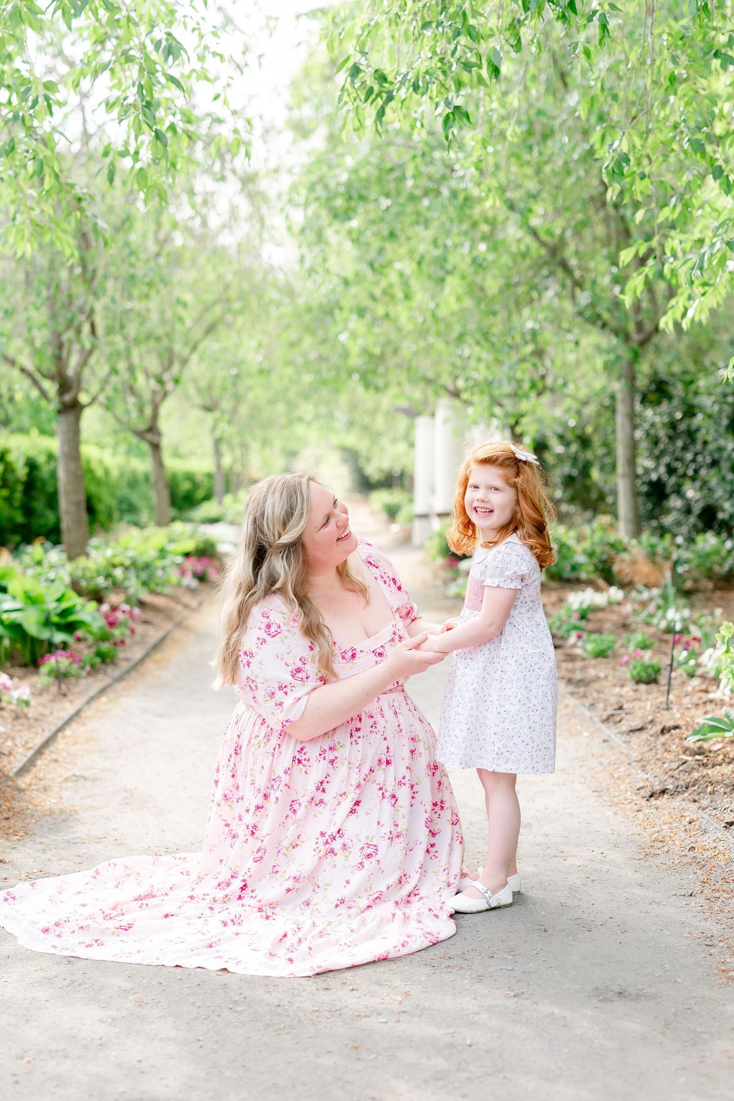 A mother in a floral dress kneels on a garden path, holding hands with her smiling red-haired daughter in a white dress.