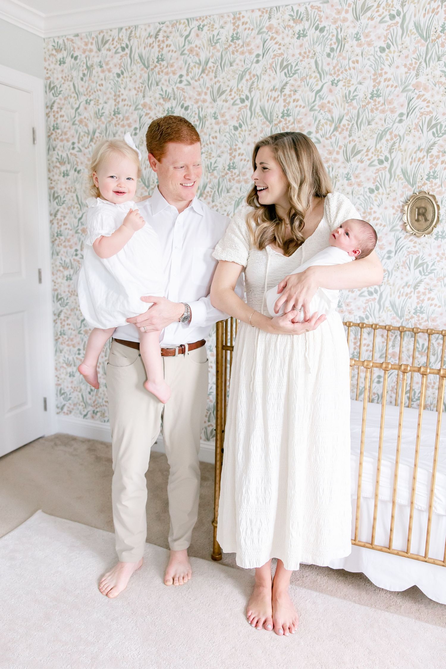 A bright nursery scene with floral wallpaper features a family of four in coordinating white and cream outfits.