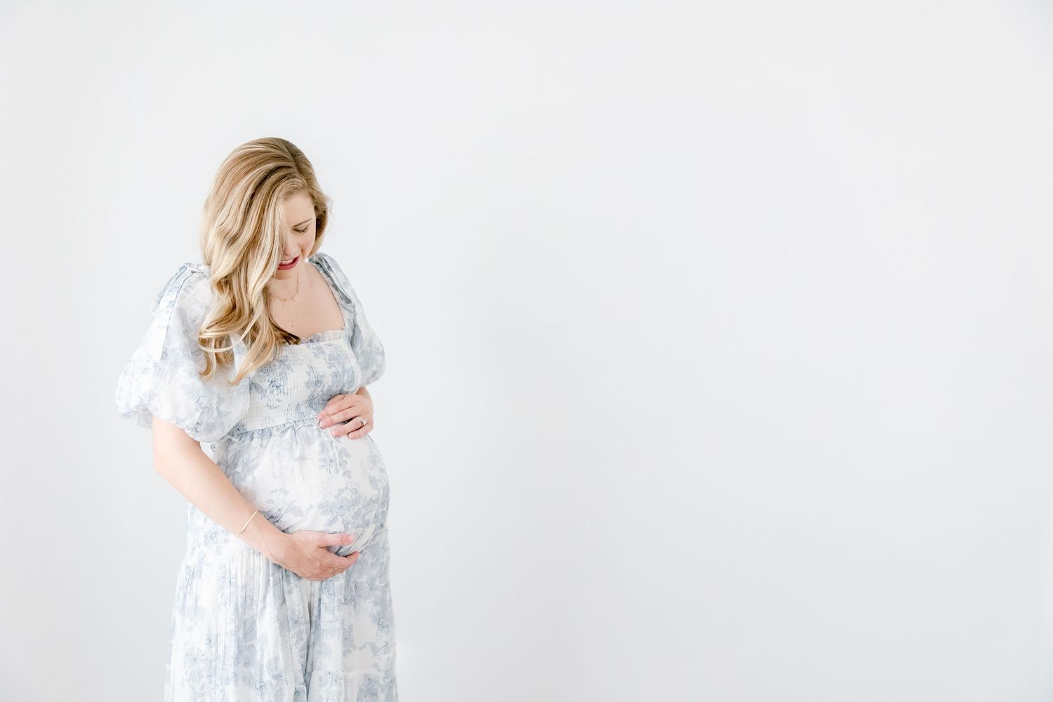 Pregnant woman in soft blue floral maternity dress poses in front of white backdrop in a maternity photo series.