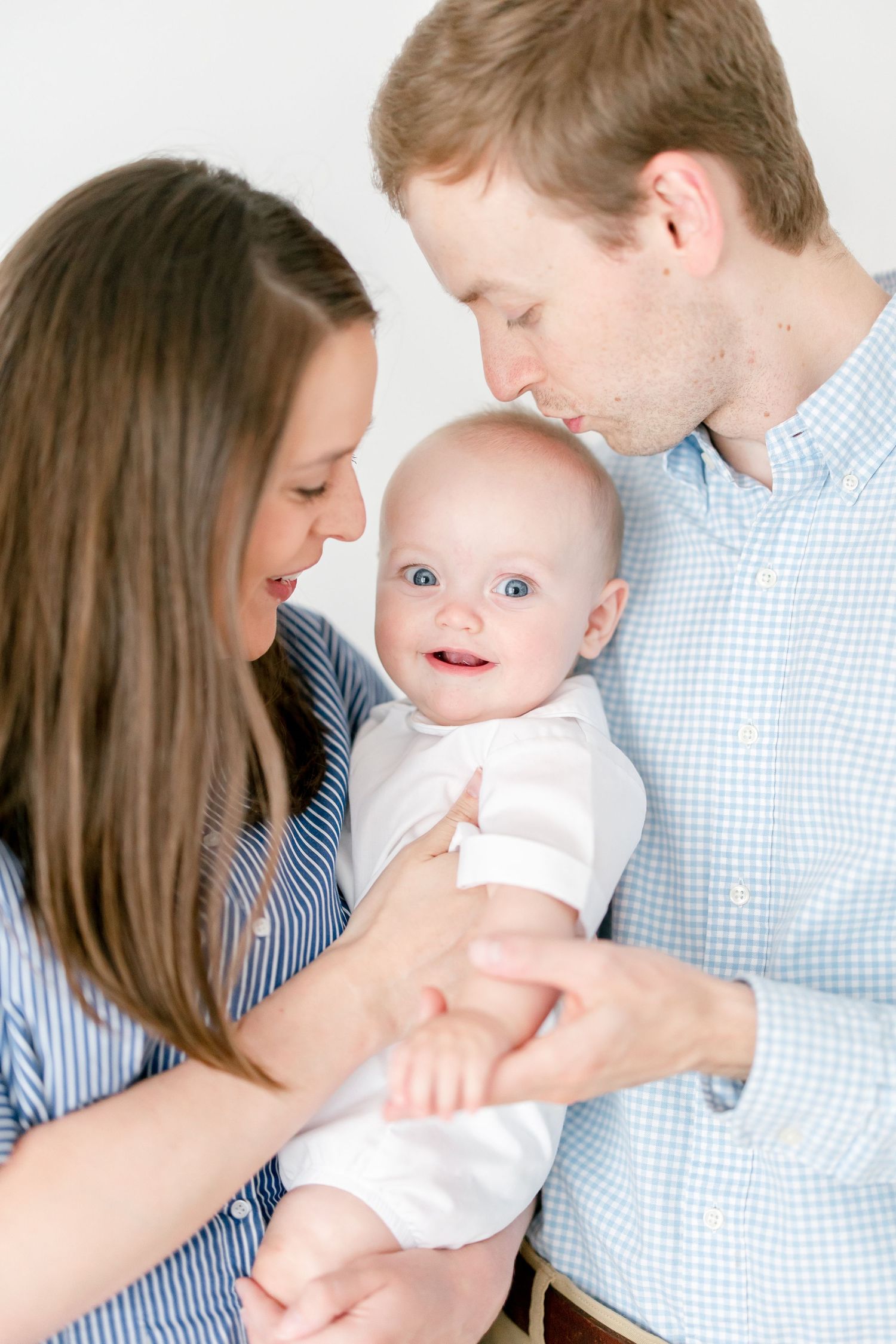 Parents in blue striped shirts snuggle close with their smiling baby in white.