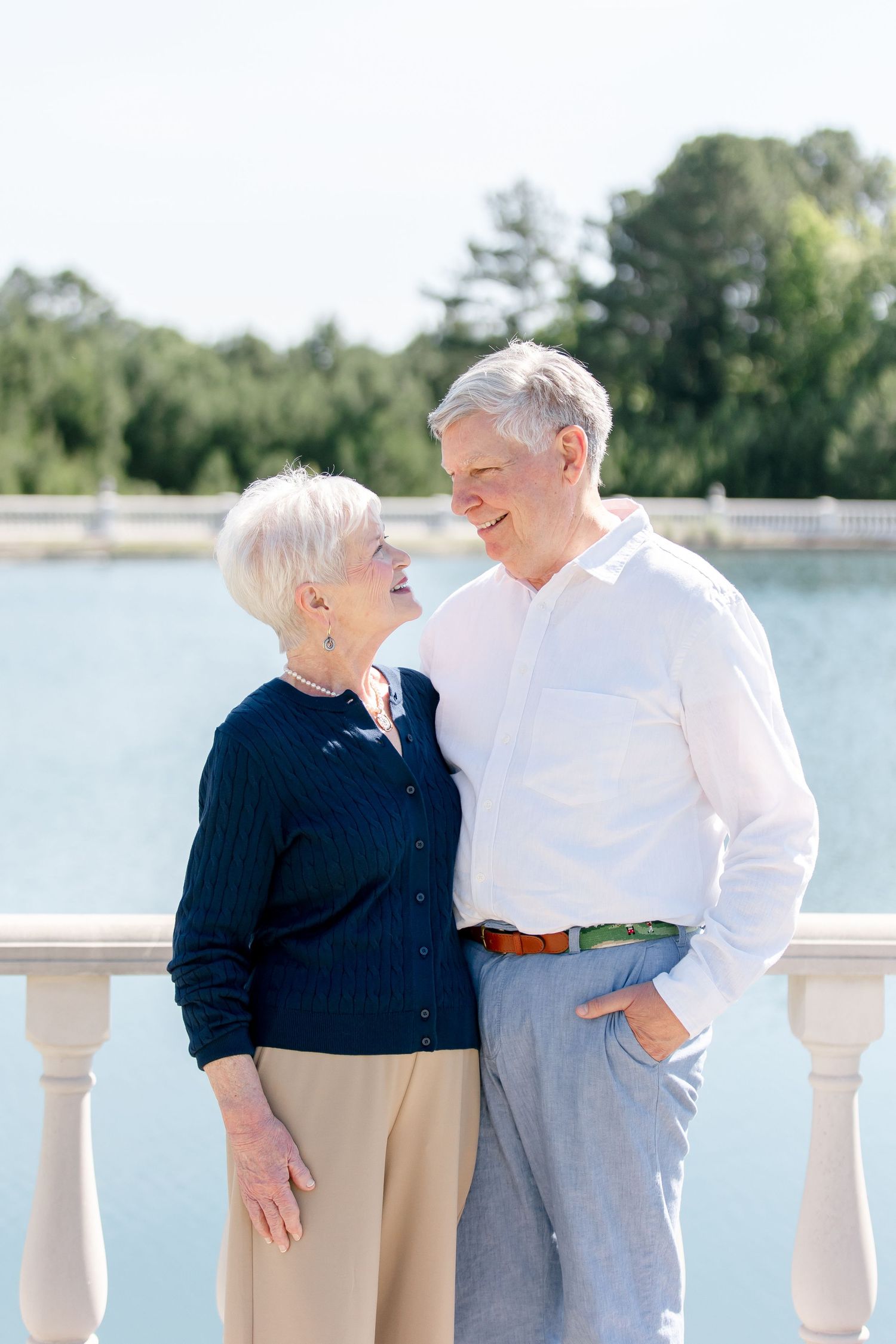 Senior couple in casual attire laughing together on waterfront balcony.