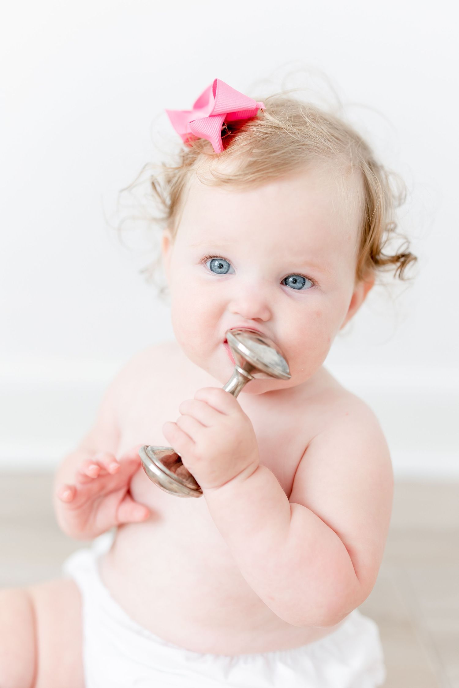 Baby with bright blue eyes and pink bow wearing white against light backdrop holds silver spoons in artistic photo series.