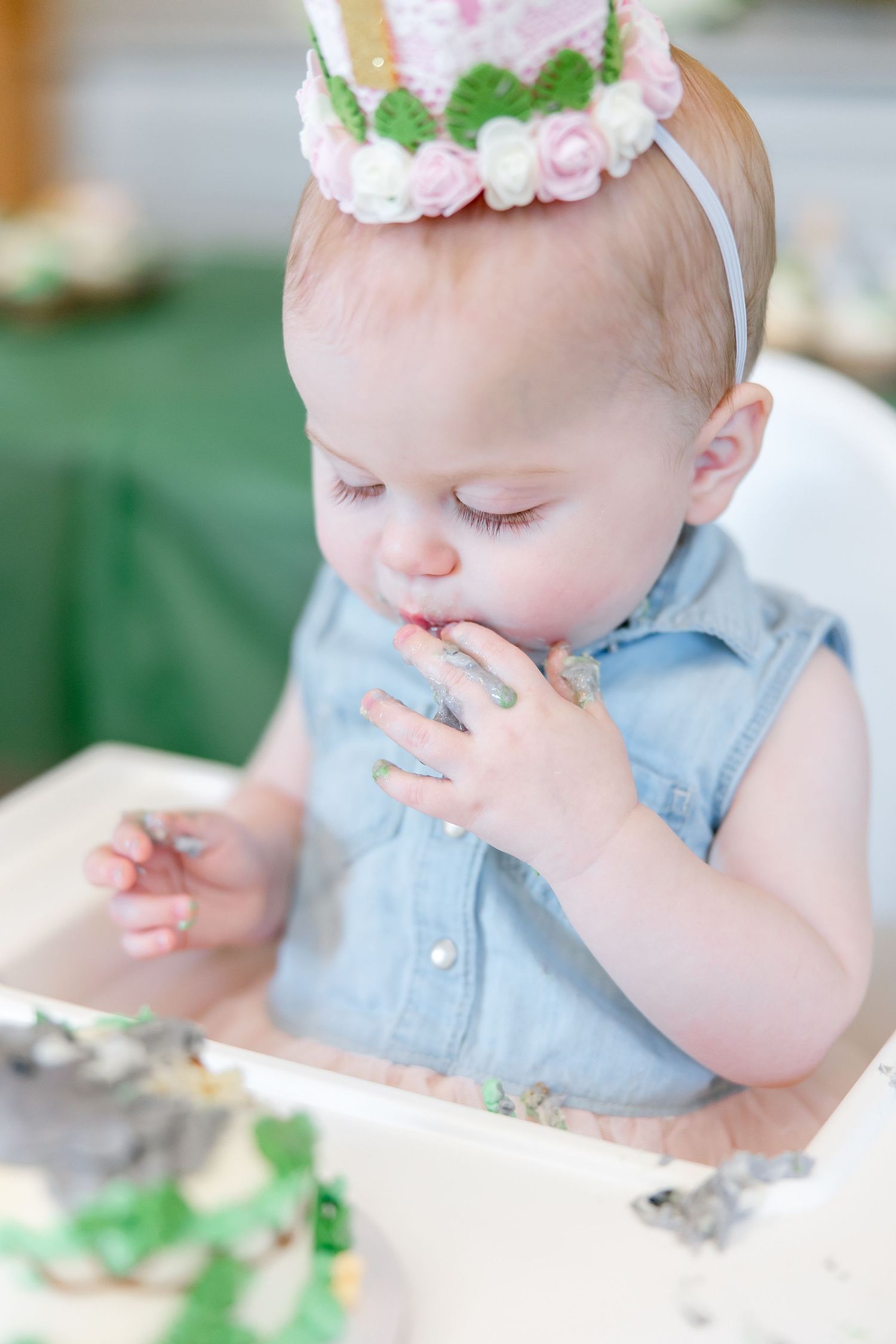 Small child wearing denim and floral crown tasting birthday cake with messy hands.