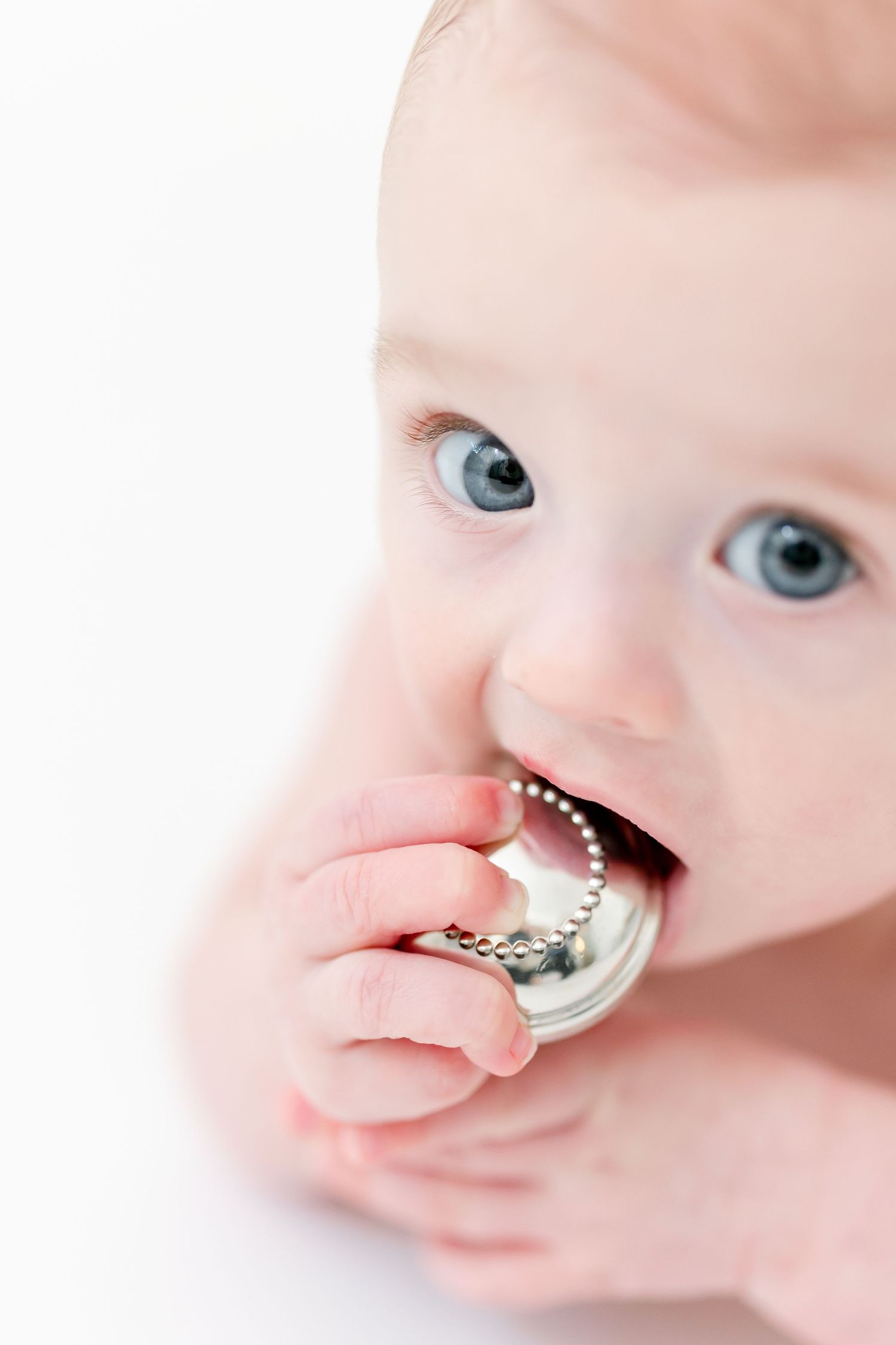 Blue-eyed baby looking up at the camera while chewing on a silver beaded teether against a bright white background.