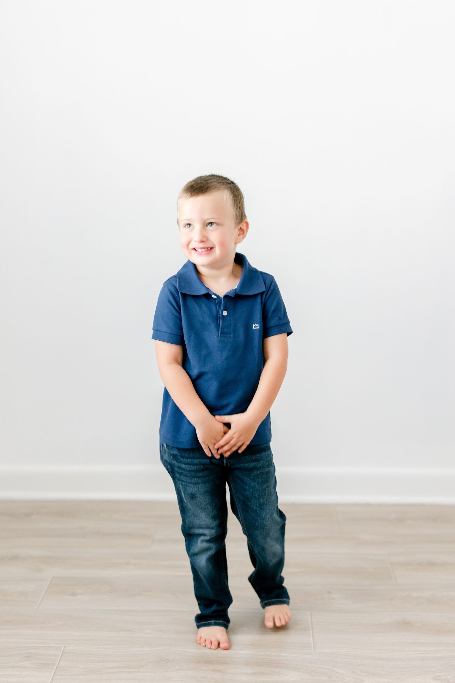Young boy in navy polo shirt and jeans plays barefoot on light wooden floors in a white room.