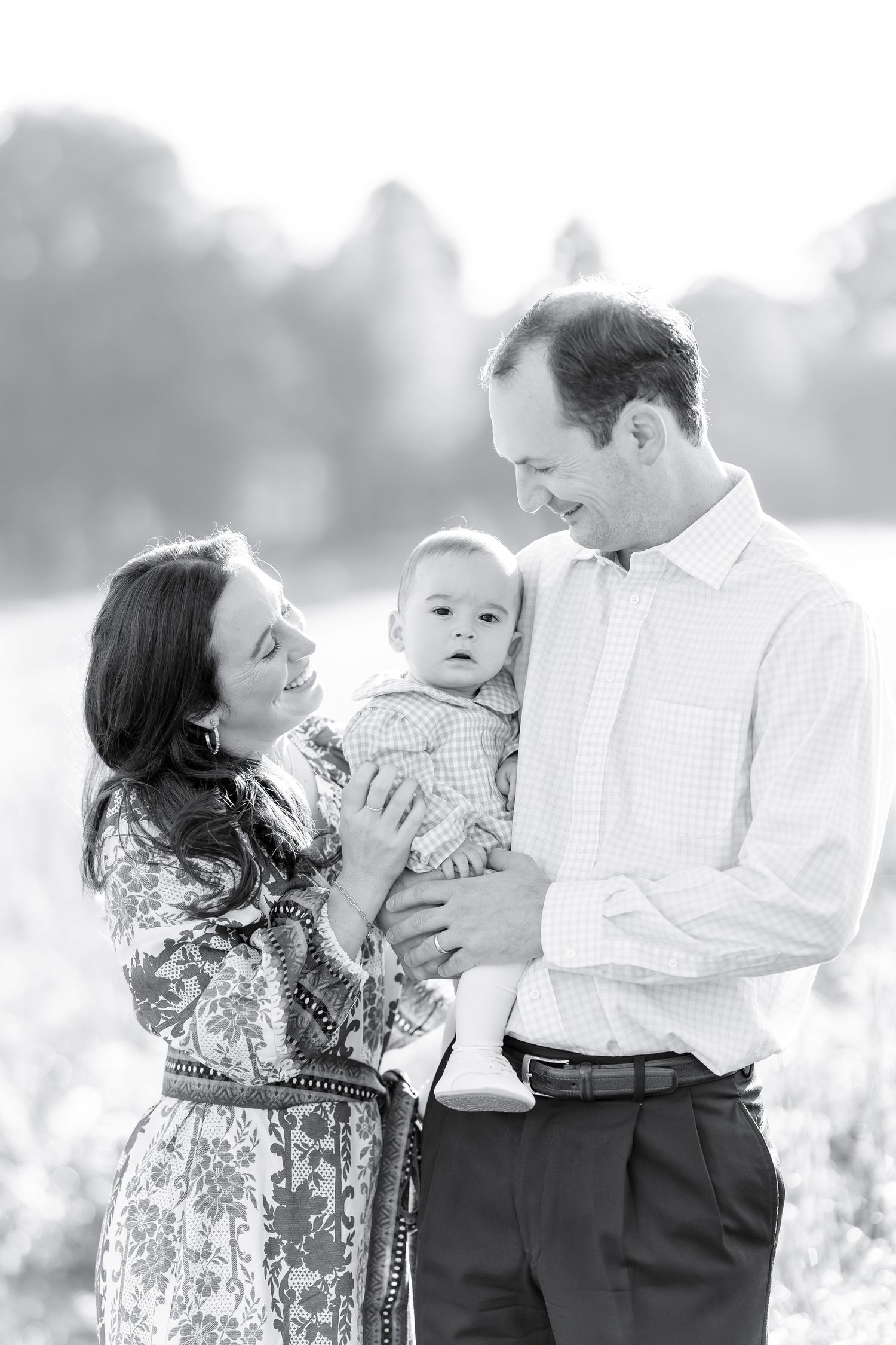 Black and white family portrait of parents smiling at baby outdoors in a sunlit field.