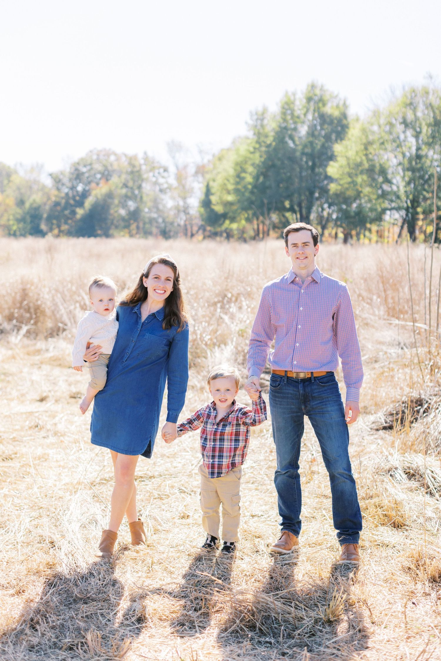 A family stands together in a sunlit field with tall golden grass and trees in the background.