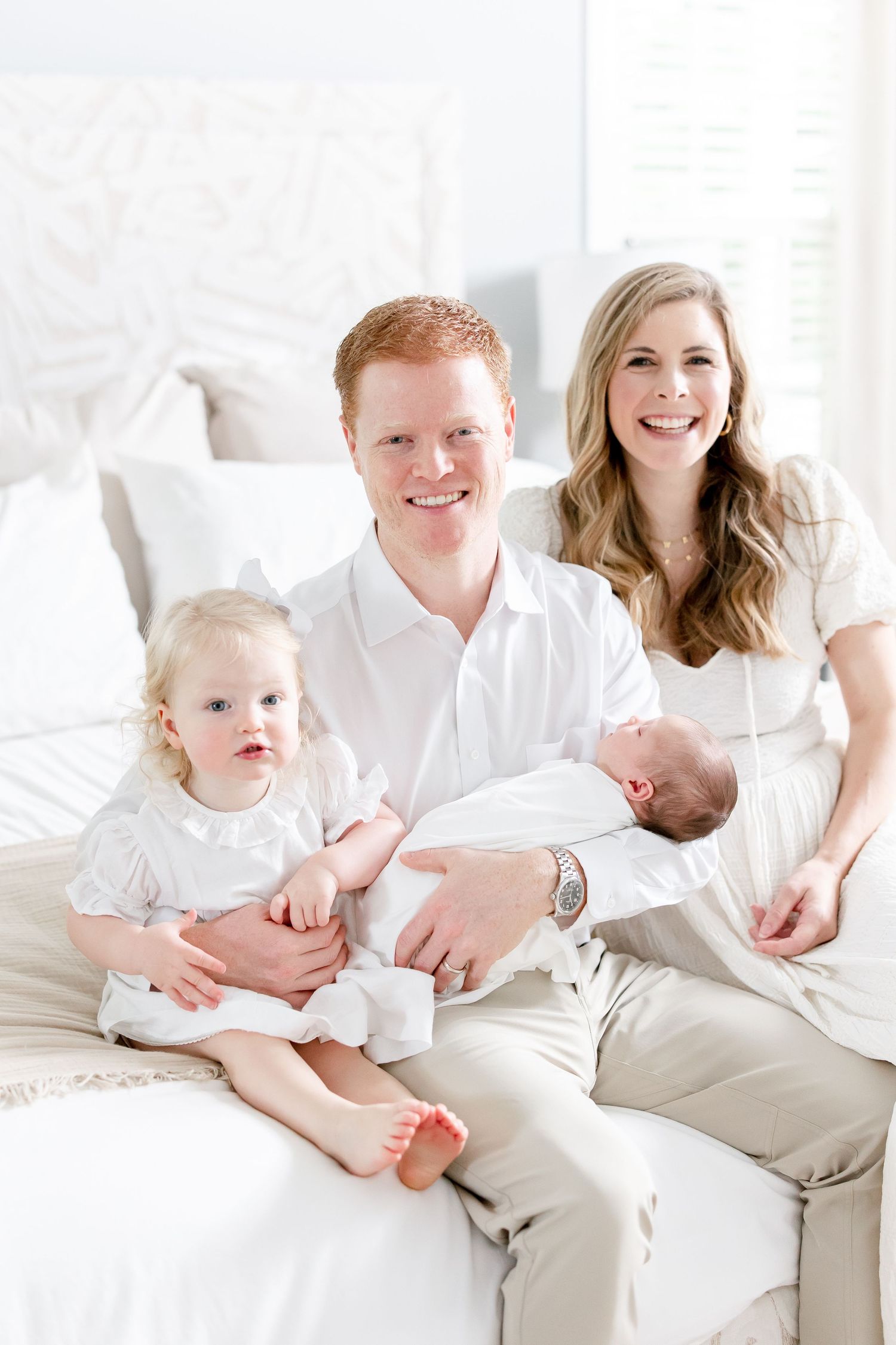 Family portrait in white outfits sitting together on bed with young children.