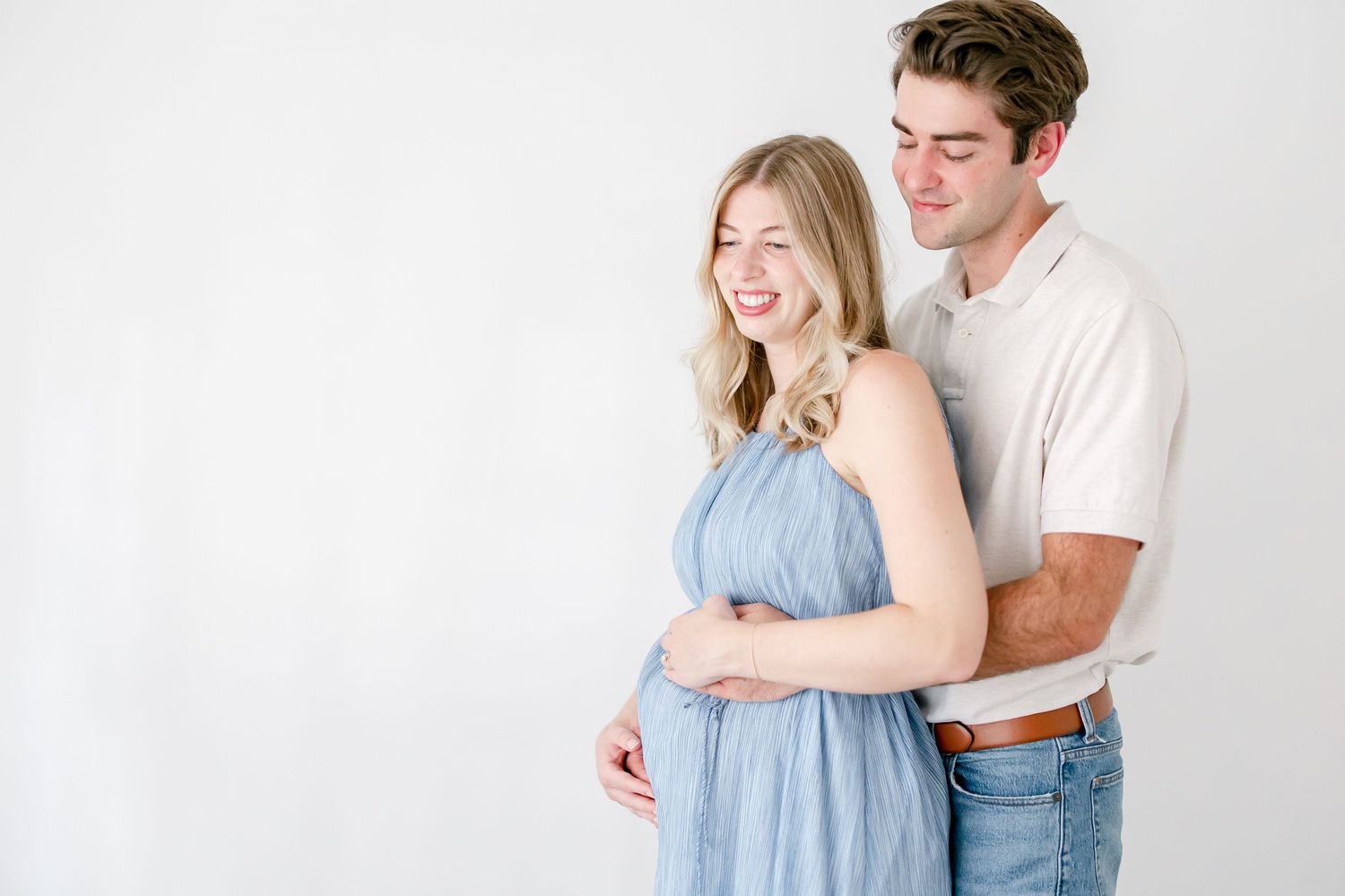 A couple embraces against a white backdrop, with her wearing a light blue dress and him in a casual white t-shirt and jeans.